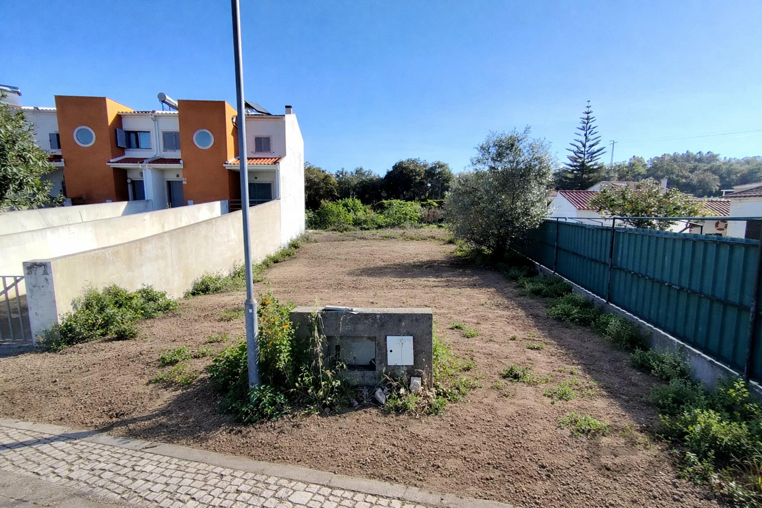 Empty dirt lot bordered by a concrete wall on the left and a blue fence on the right, with an orange townhouse under a clear blue sky