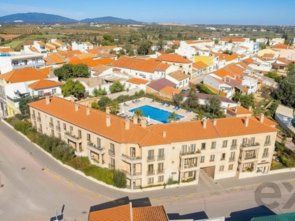 Aerial view of a sunny town with orange-tiled roofs, white buildings, and a central blue swimming pool courtyard surrounded by palm trees and loungers under a clear sky