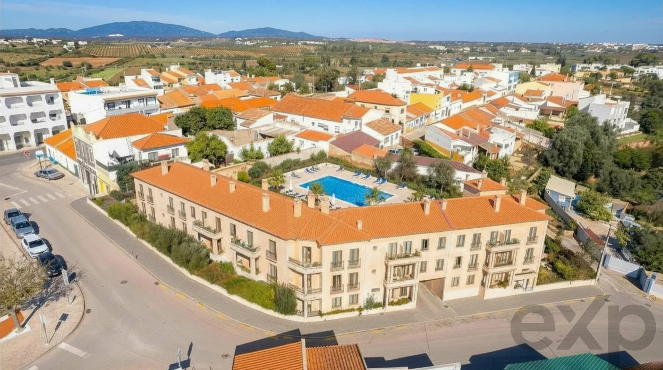 Aerial view of a sunny town with orange-tiled roofs, white buildings, and a central blue swimming pool courtyard surrounded by palm trees and loungers under a clear sky