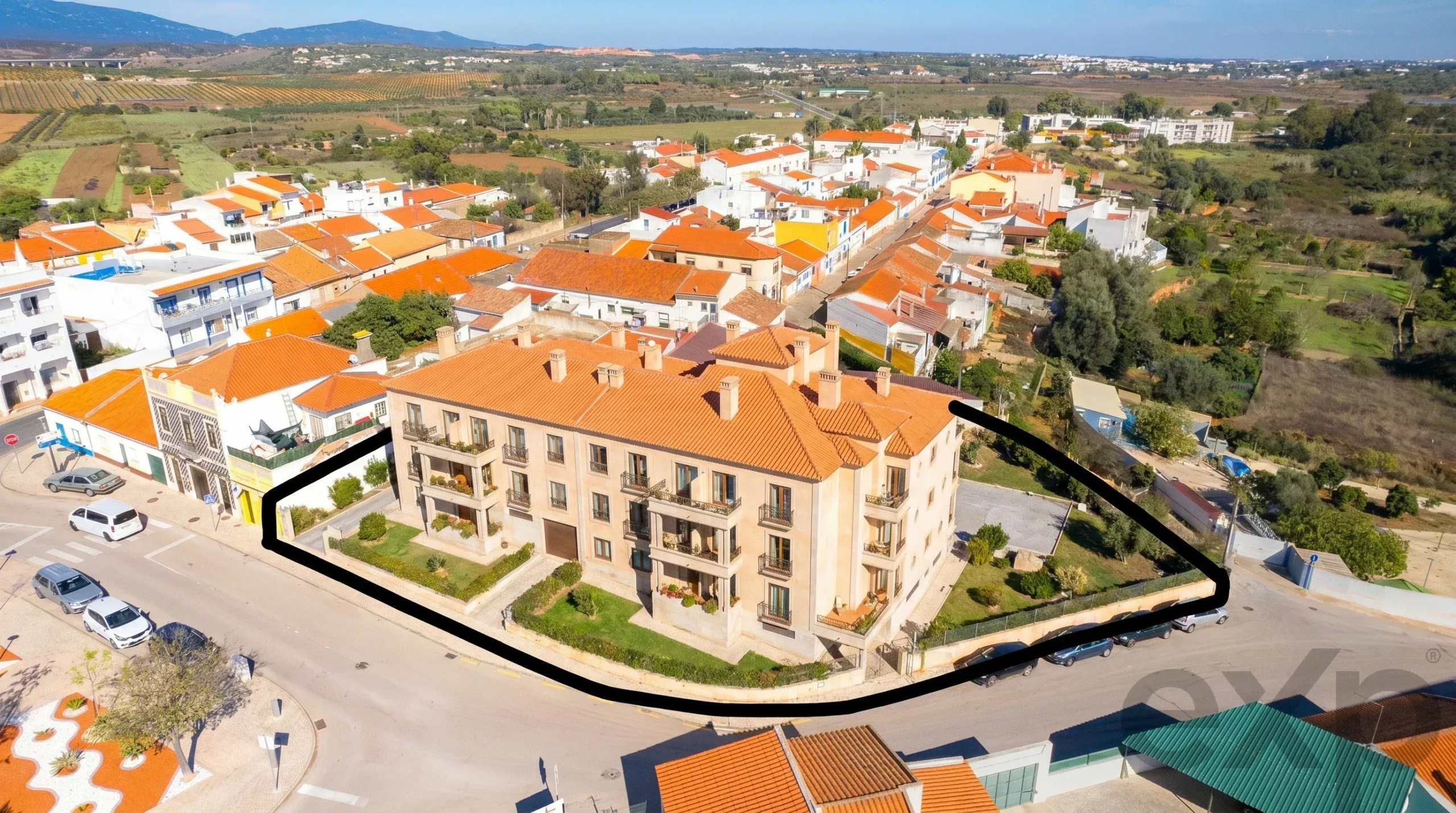 Aerial view of a town with orange tiled roofs; a large beige apartment building sits at a street corner.
