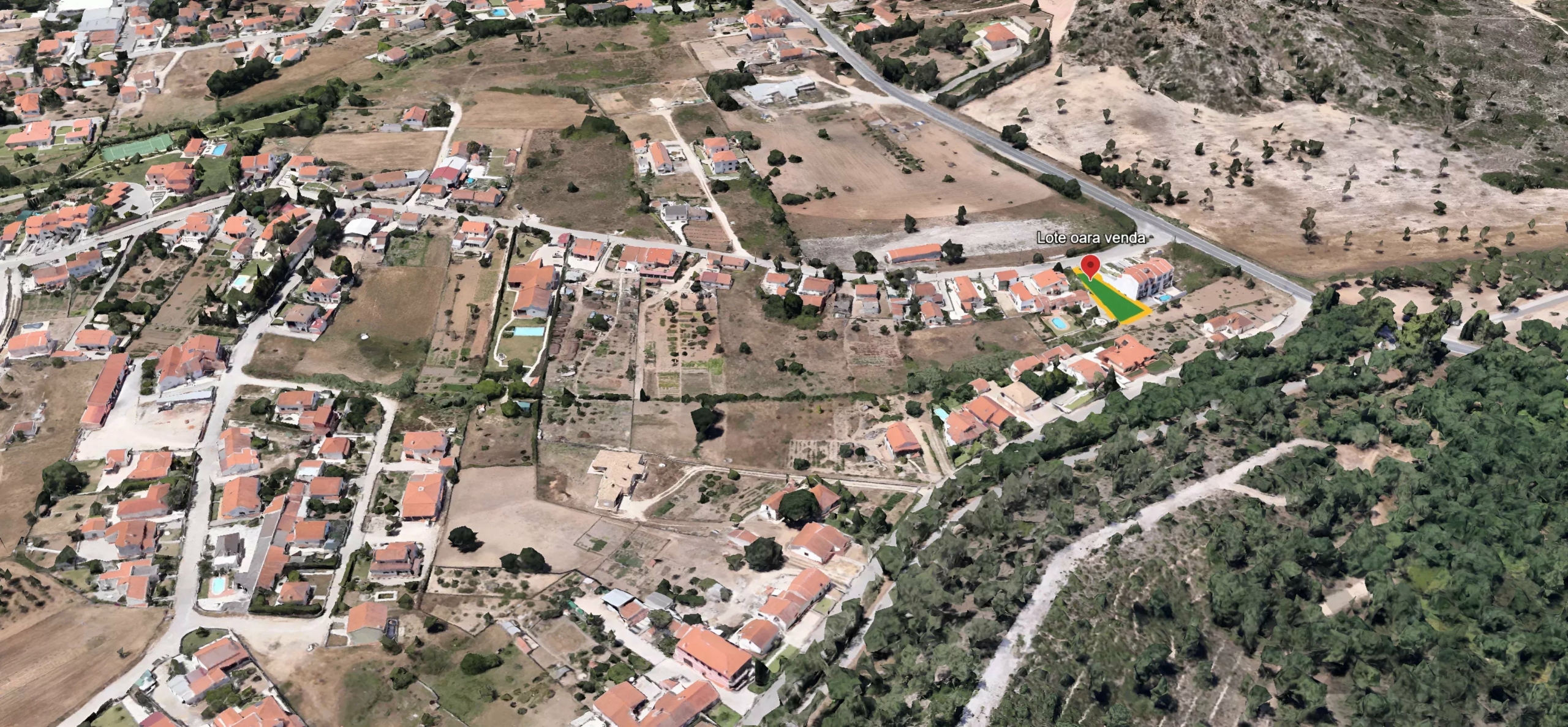 Aerial view of a rural town with red-roofed houses and fields; a highlighted triangular land parcel for sale beside a road.