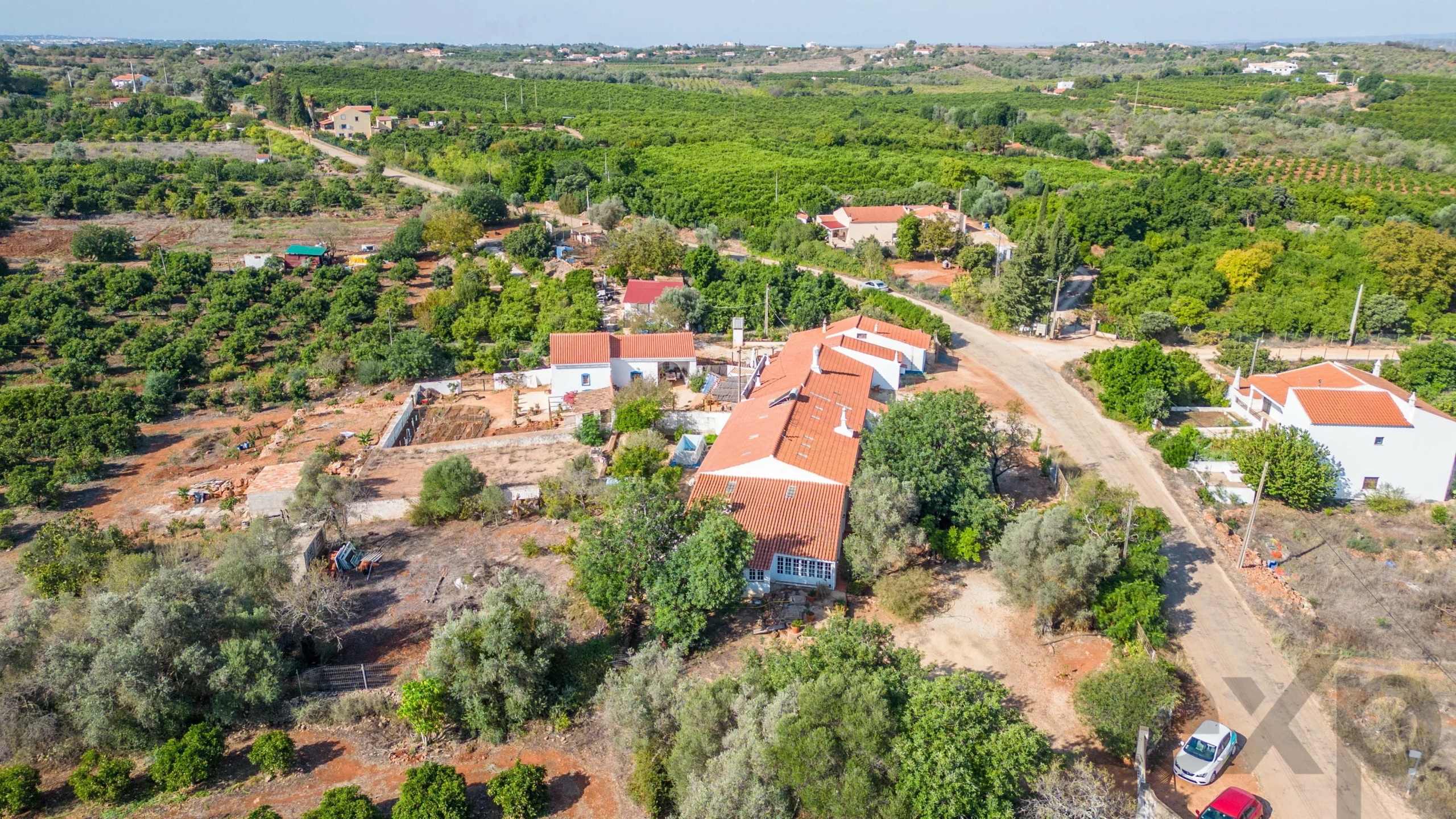 Aerial view of a rural village with white houses and red-tiled roofs among olive trees and green fields.