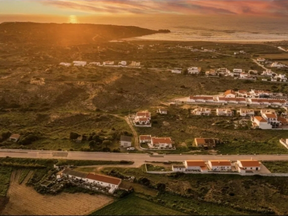 Aerial view of a coastal village at sunset, with white houses and red-tiled roofs along a road toward the sea.