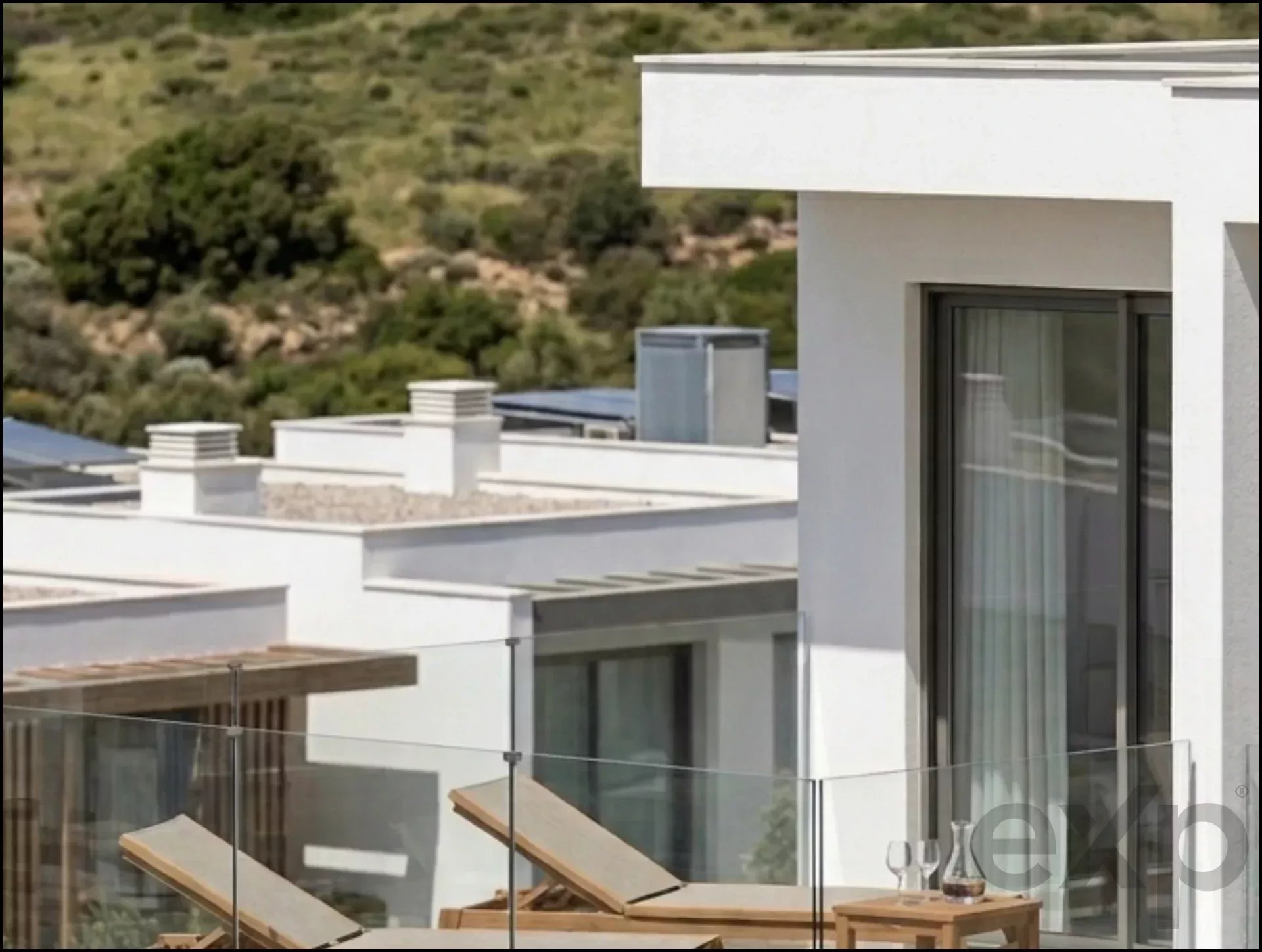Modern white contemporary home rooftop terrace with glass railing, wooden sun loungers, and a glass dining table set against a hillside landscape.