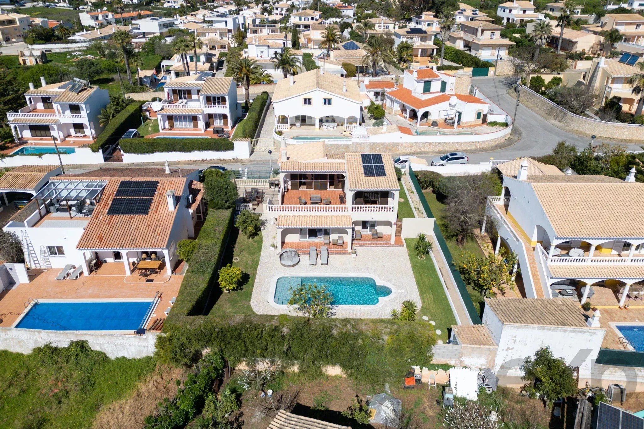 Aerial view of a sunny Mediterranean-style neighborhood with white houses, tiled roofs, palm trees, and several rectangular swimming pools in backyards.