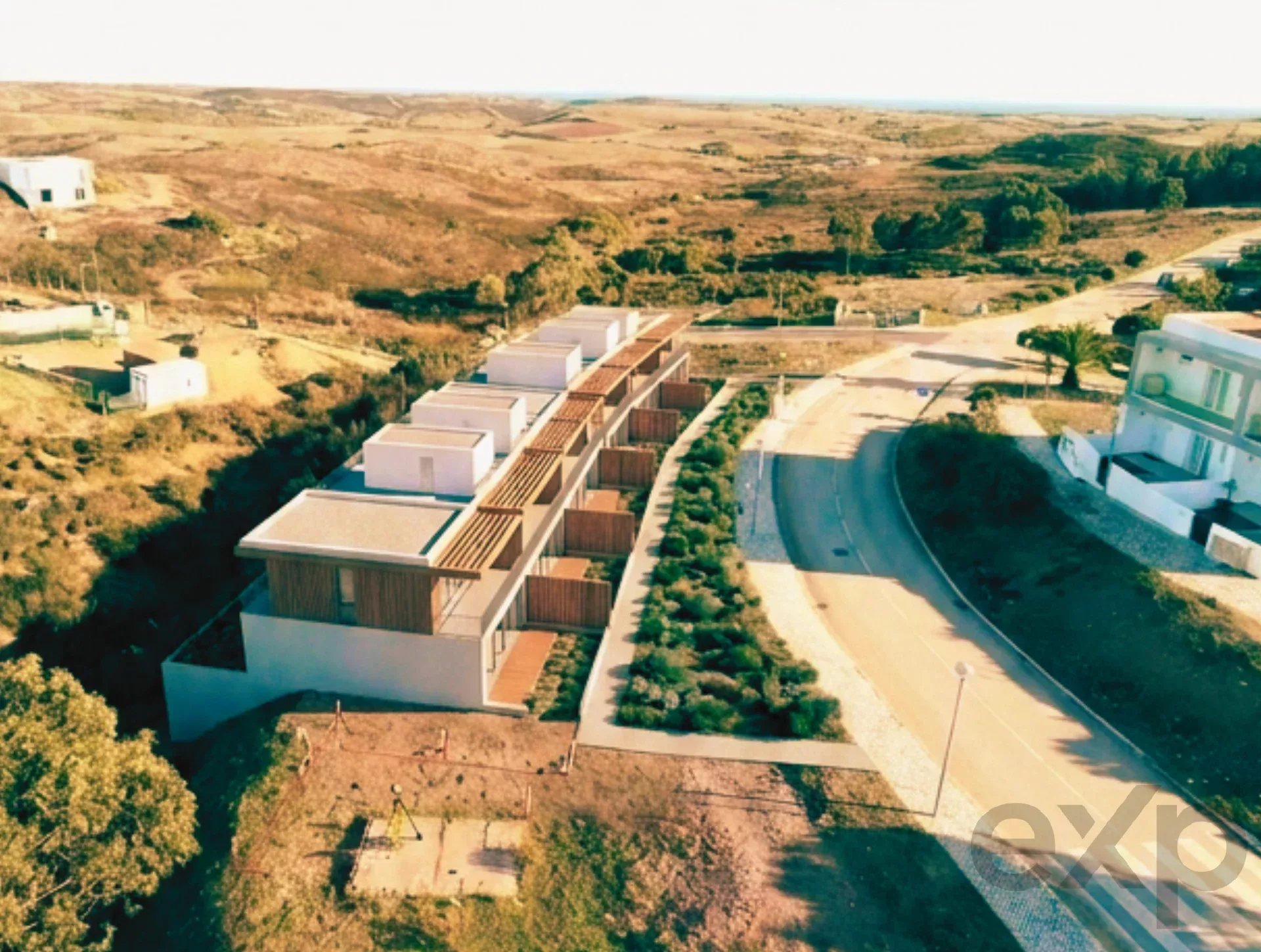 Aerial view of modern white townhouses along a curved road in a dry, hilly landscape with sparse vegetation.