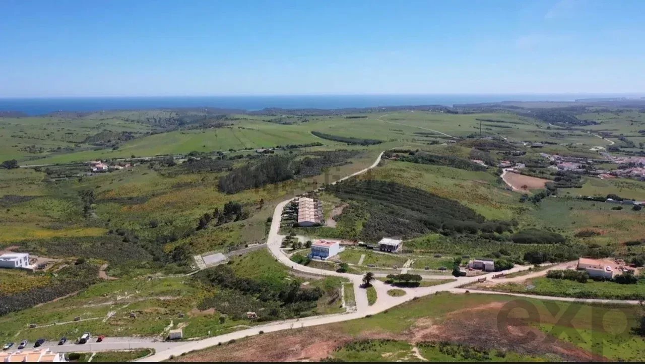 Aerial view of rolling green hills dotted with houses, winding roads, and a distant blue sea on the horizon.