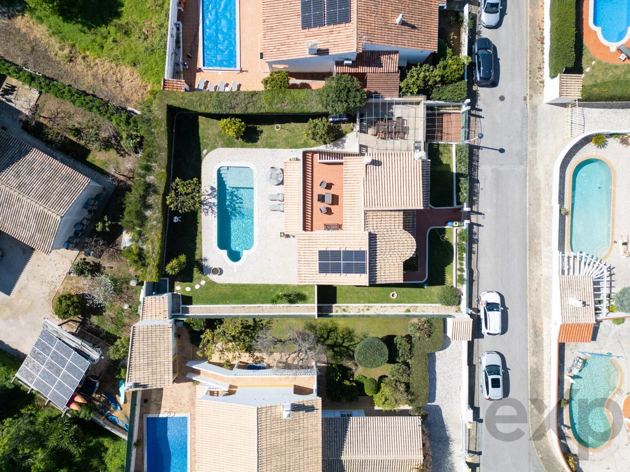 Aerial view of a suburban neighborhood showing terracotta-tiled roofs, backyards with rectangular blue pools, and solar panels on some roofs with cars along a street on the right.