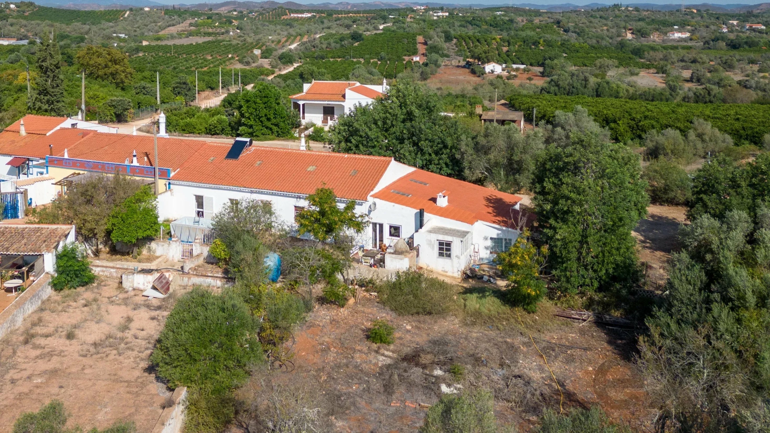Aerial view of a rural hillside village with white houses and red-tiled roofs surrounded by olive trees and cultivated fields on terraced slopes in the distance.