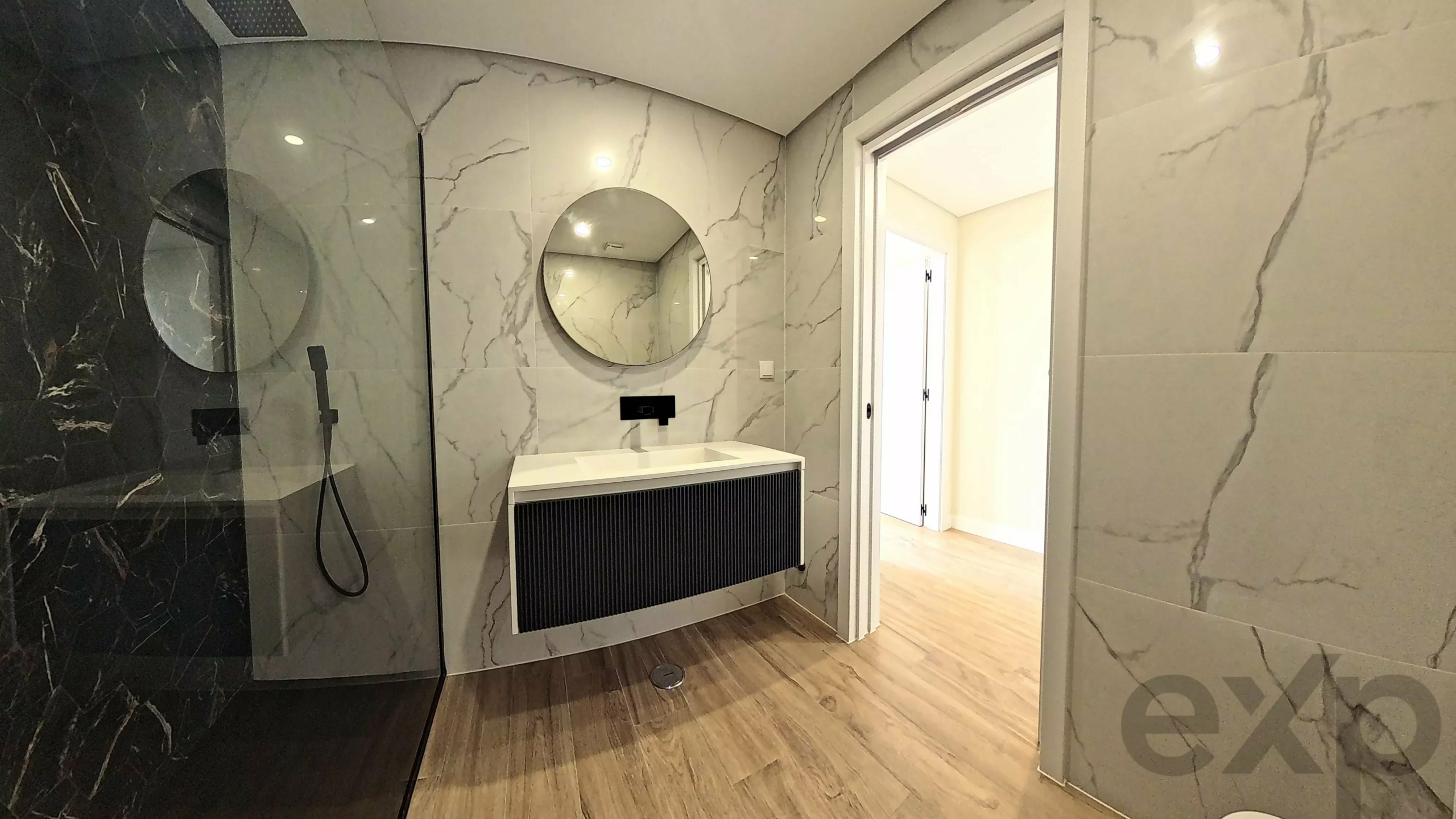 Modern bathroom with marble walls, round mirror above a white sink, black floating vanity, and glass shower on the left.