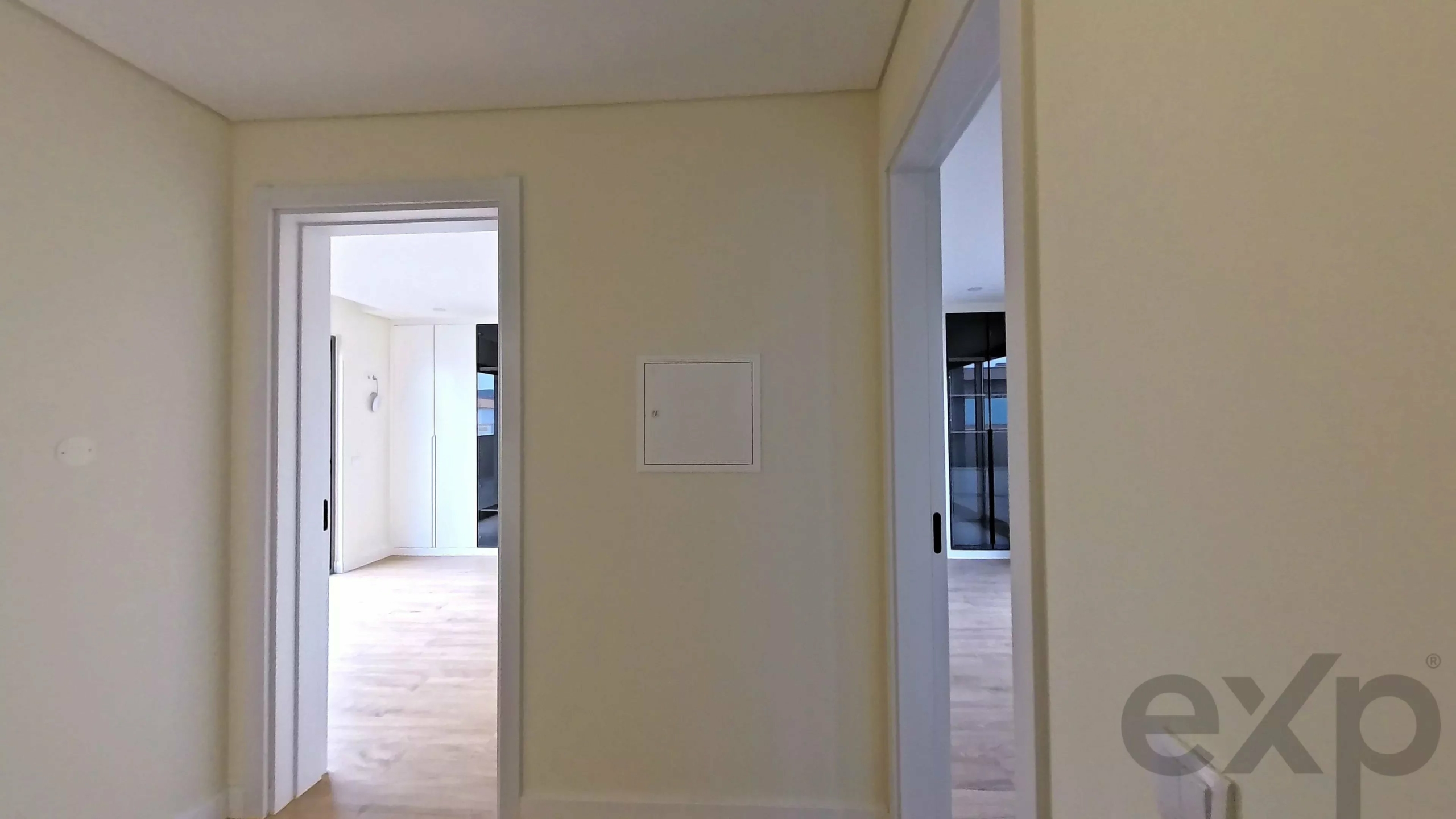 Interior hallway with two doorways to adjacent rooms; view into a bright kitchen or living area through the left doorway, and a windowed room through the right doorway; a small wall access panel is centered on the wall.
