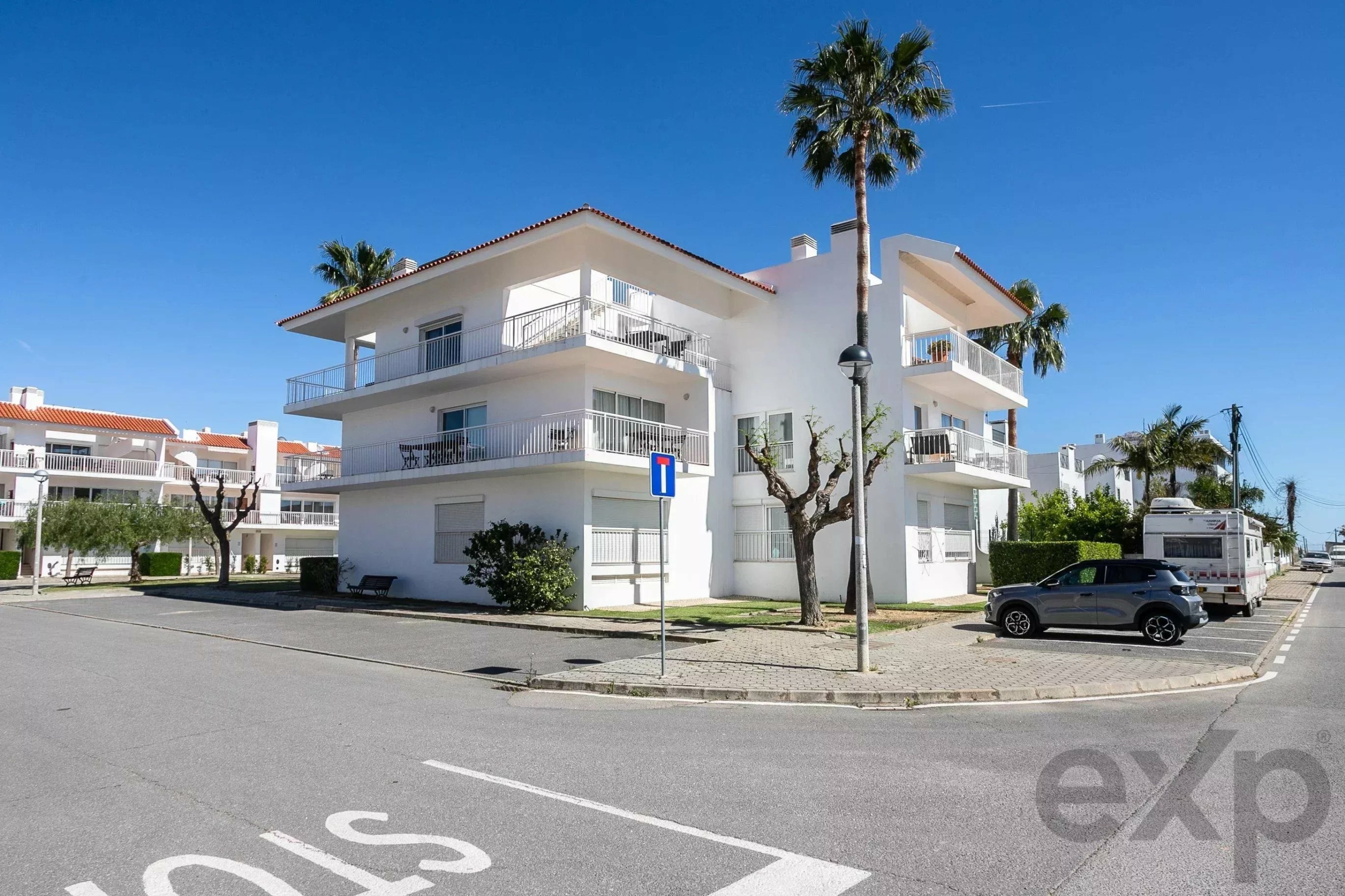 White three-story apartment building with balconies under a clear blue sky, palm trees, and parked cars along the street.