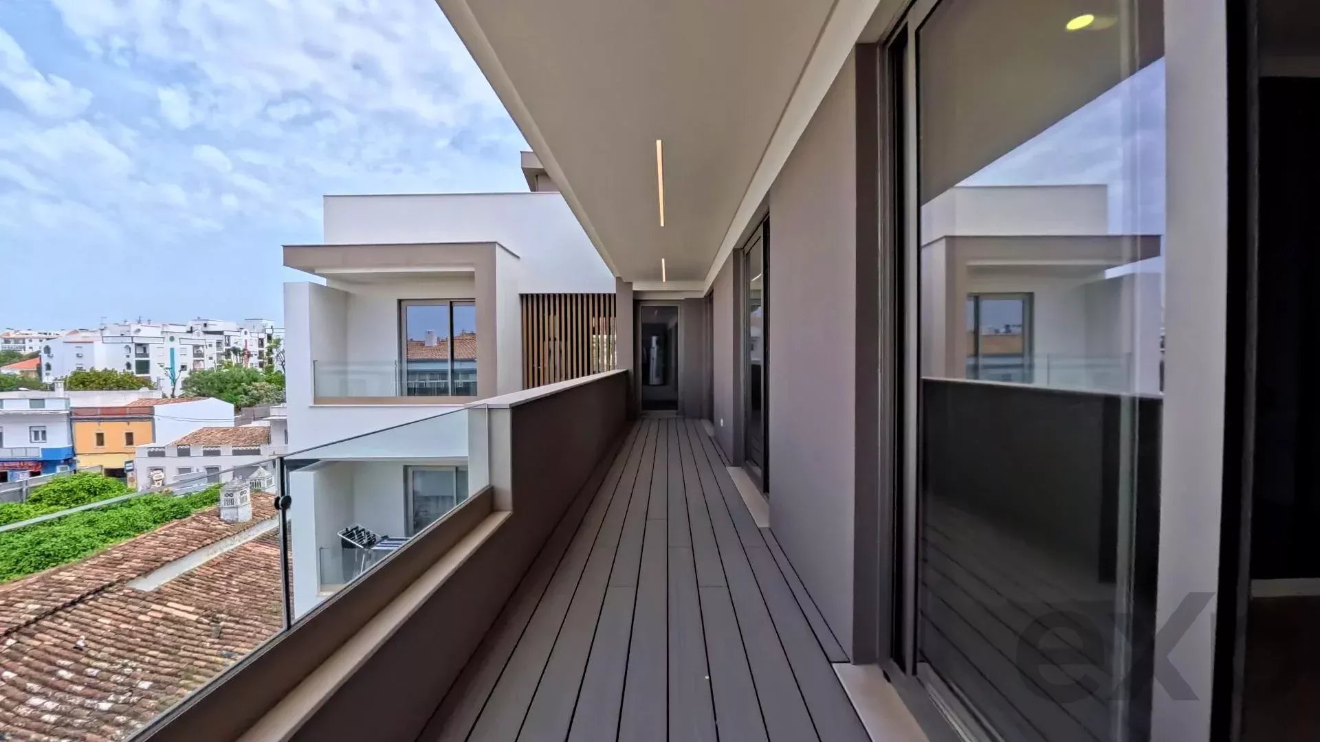 Long outdoor balcony of a modern building with gray decking, glass railing, and sliding glass doors, city neighborhood beyond.