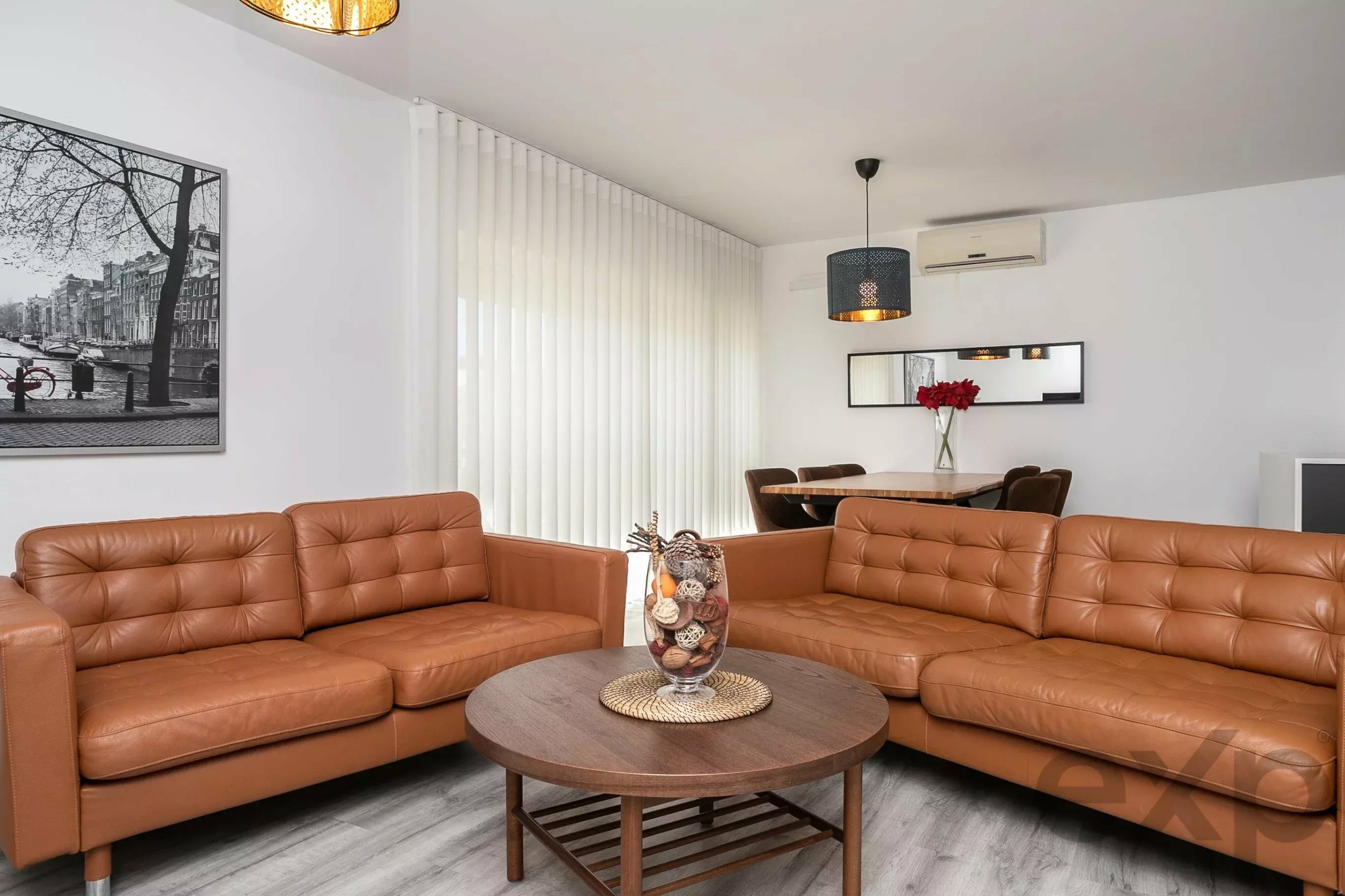 Living room with tan leather sectional sofa, round wooden coffee table, and vertical white blinds.