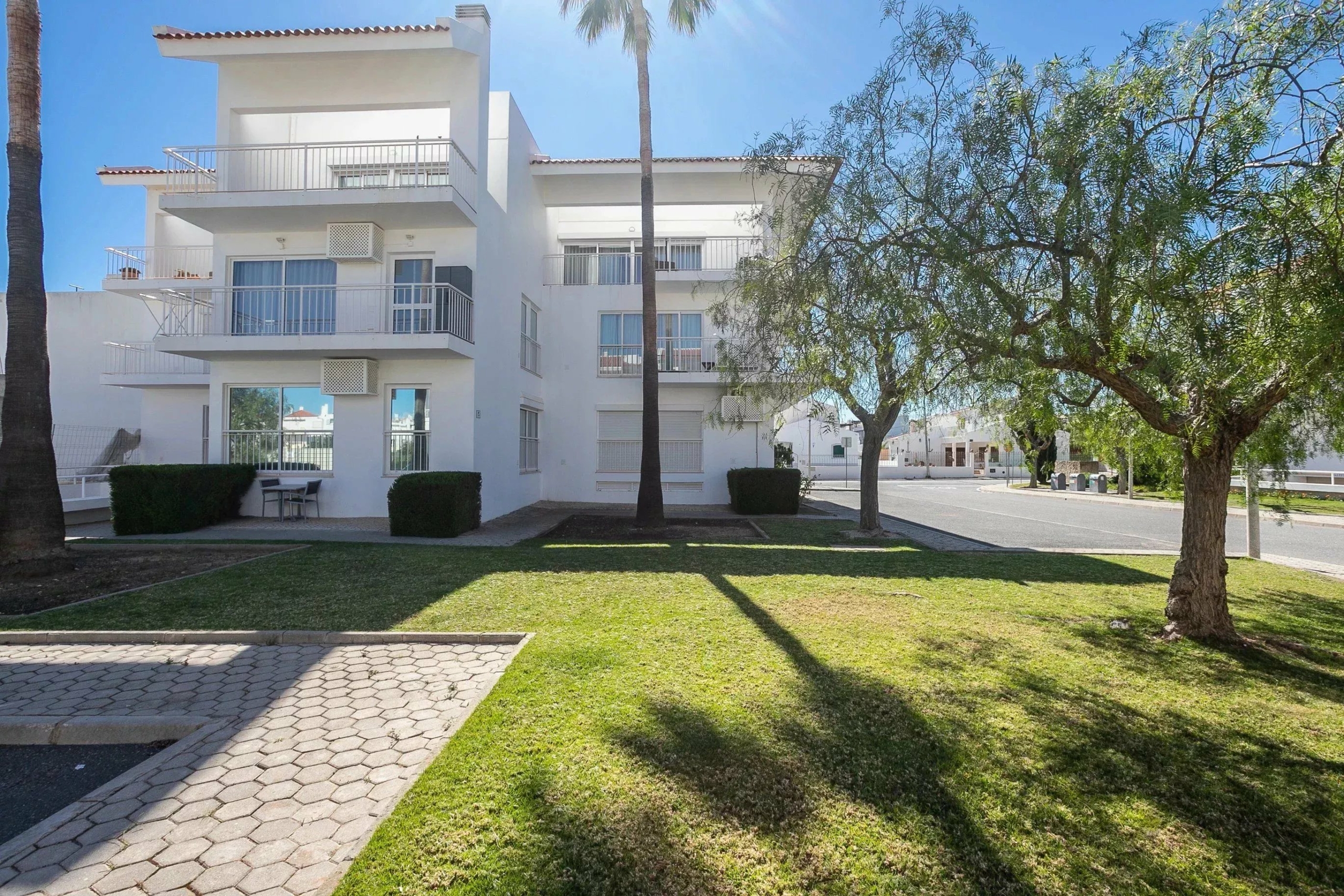 White, multi-story apartment building with balconies, set in a sunny courtyard with palm trees and a grassy lawn.