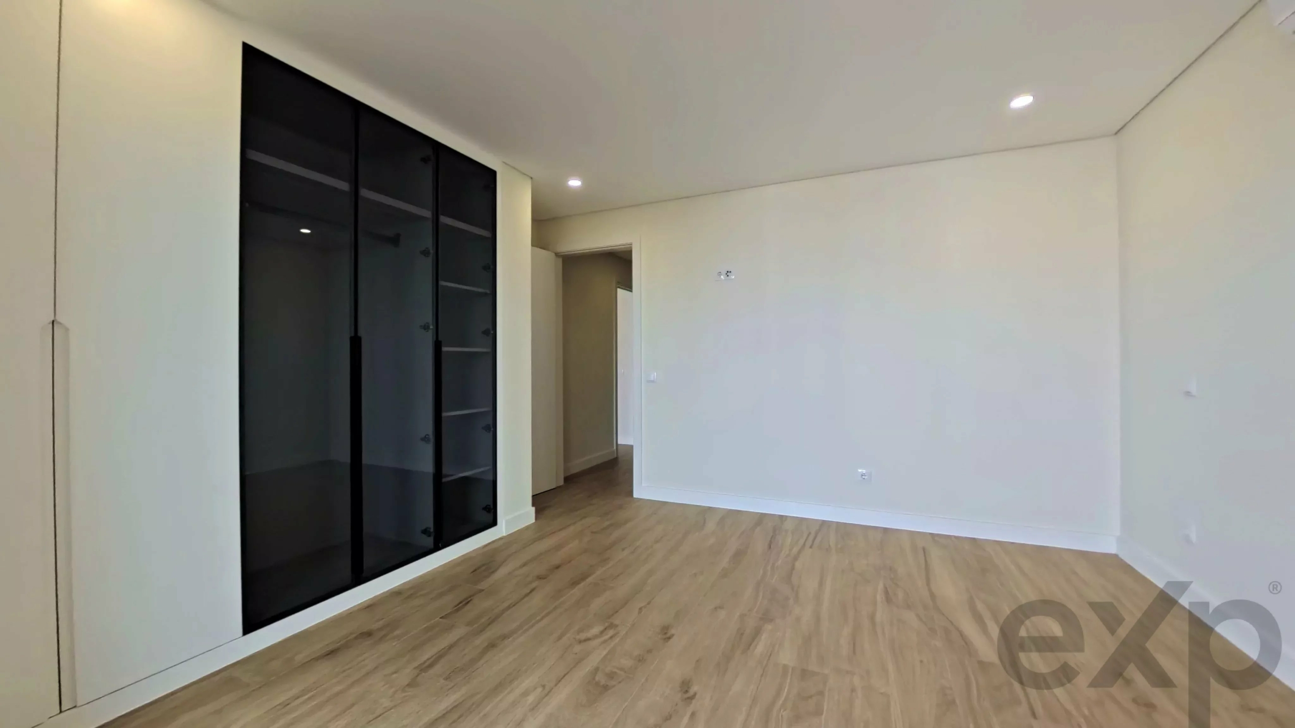 Empty, unfurnished room with light wood floors, white walls, recessed ceiling lights, and a tall black glass-front storage cabinet along the left wall.