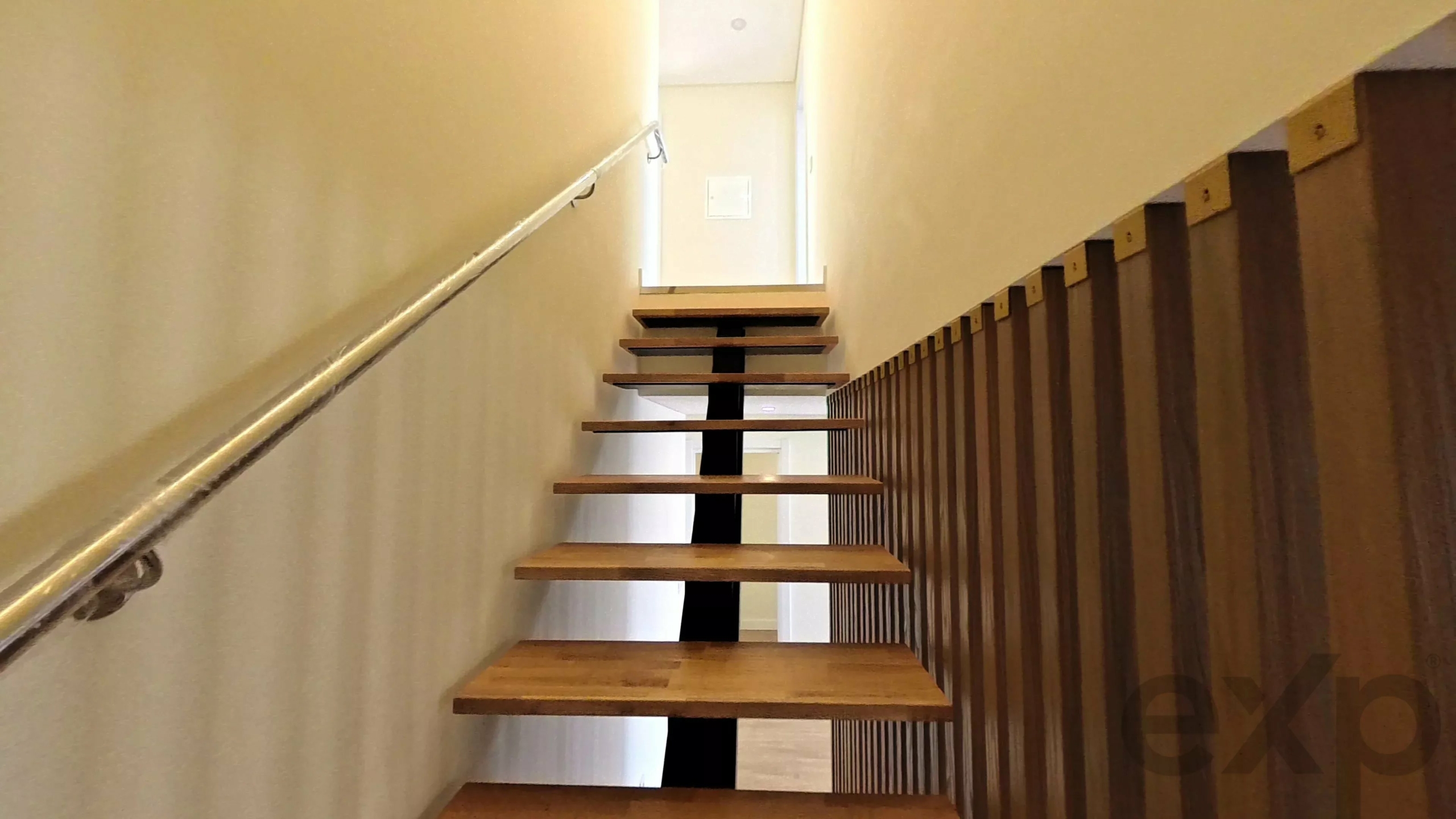 Low-angle view of a narrow indoor staircase with floating wooden steps, a metal handrail on the left, and a vertical wooden slat railing on the right leading upward.