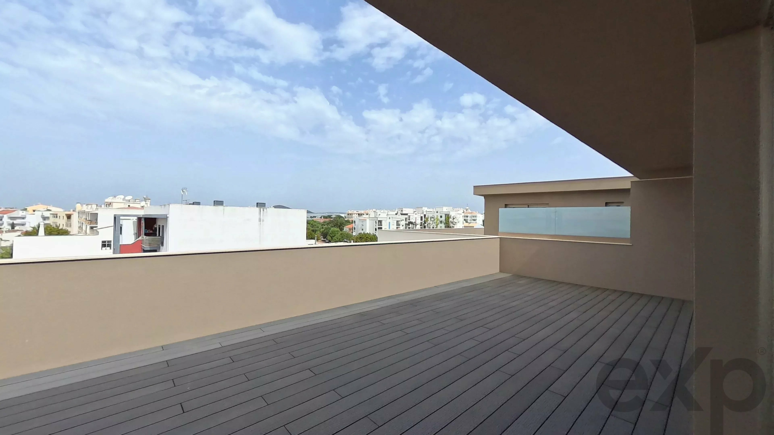 Rooftop terrace with dark wooden deck and beige parapet, overlooking a cityscape under a blue sky with scattered clouds.