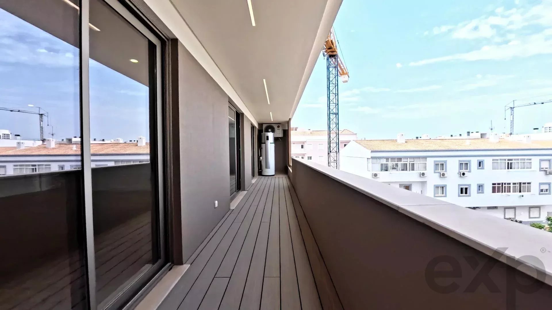 Long apartment balcony with sliding glass doors and wooden deck, railing on the right, construction crane visible against a blue sky.