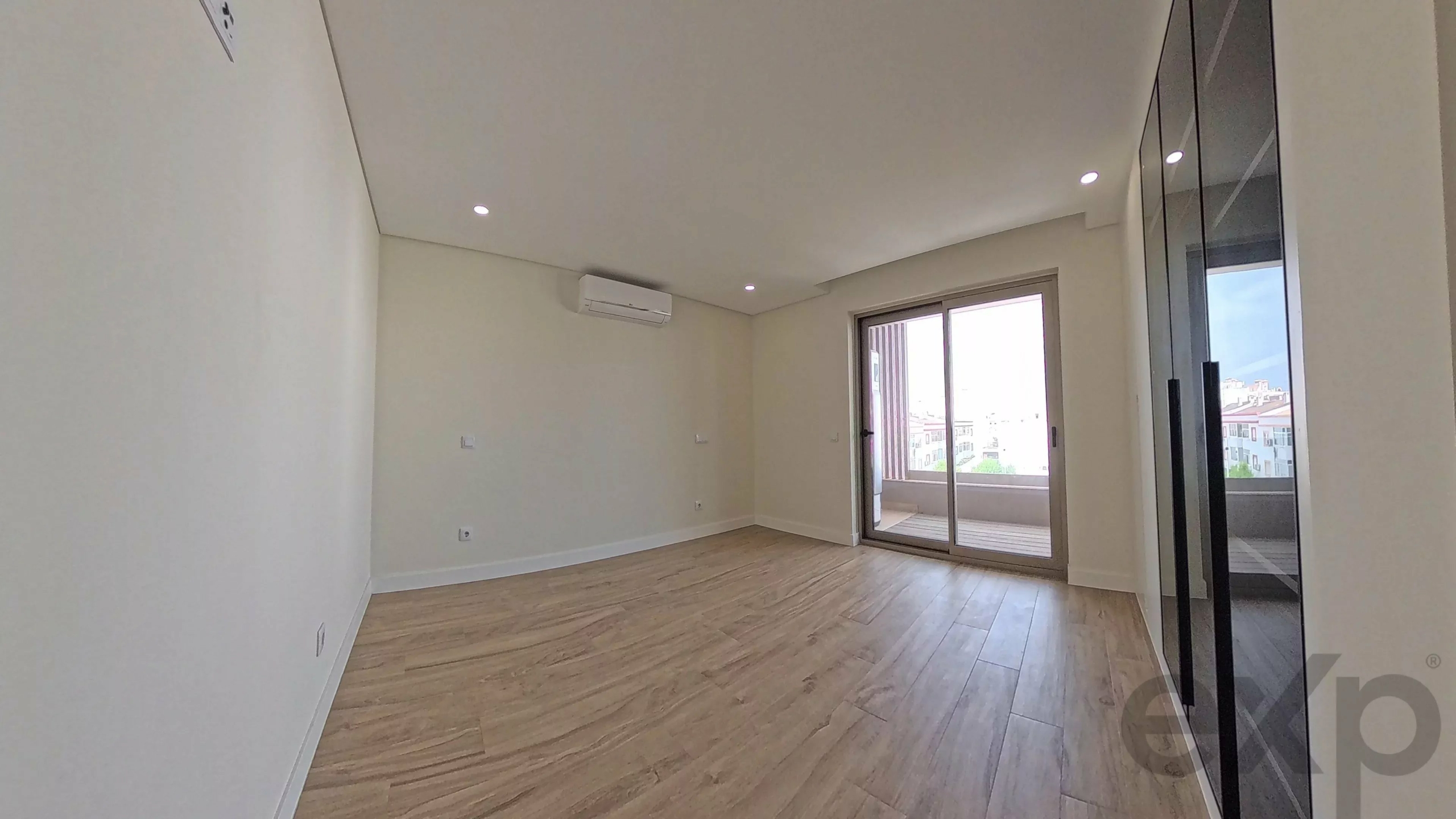 Empty living room with light wood flooring, white walls, a wall AC unit, and sliding glass doors to a balcony with a city view