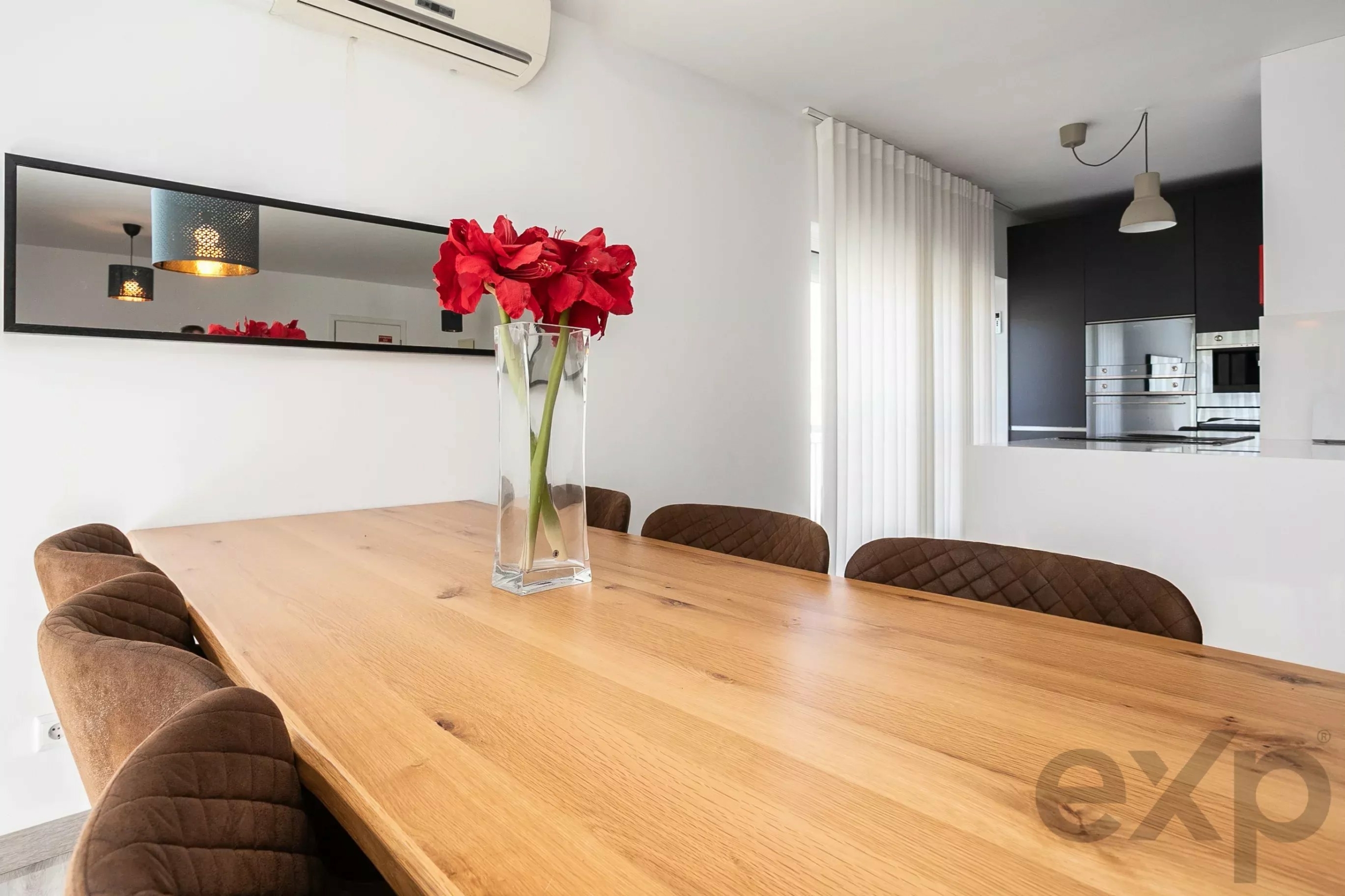 Red amaryllis flowers in a tall glass vase on a wooden dining table in a bright modern kitchen/dining area with brown quilted chairs.