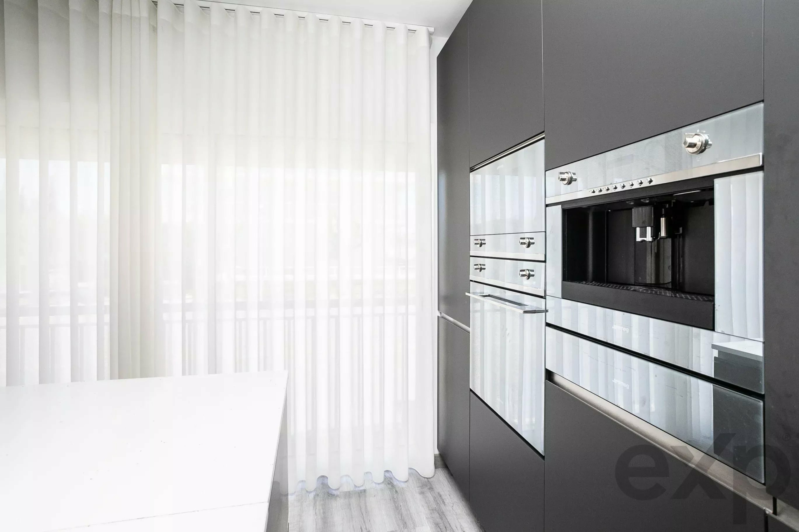 Modern kitchen with stainless steel built-in ovens and dark gray cabinetry on the right, white sheer curtains on the left.