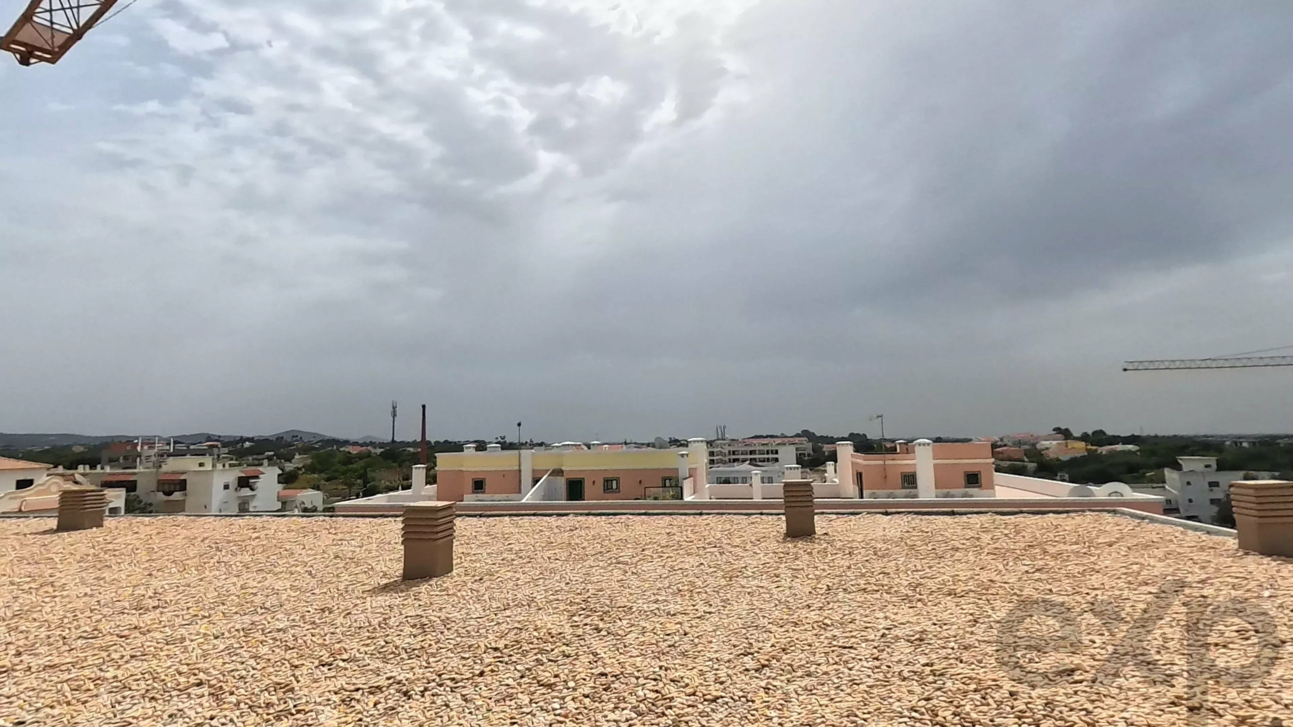 Flat gravel rooftop in foreground with chimneys, city buildings on the horizon, under a cloudy sky