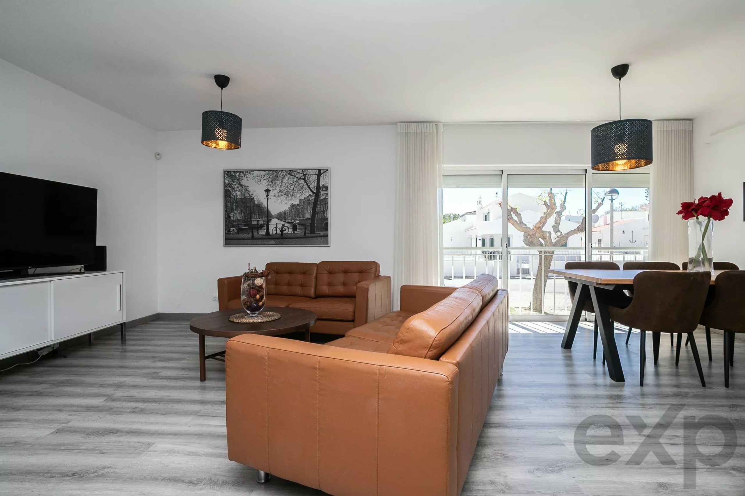 Modern living room with brown leather sofas, round coffee table, and a wall-mounted TV on the left.