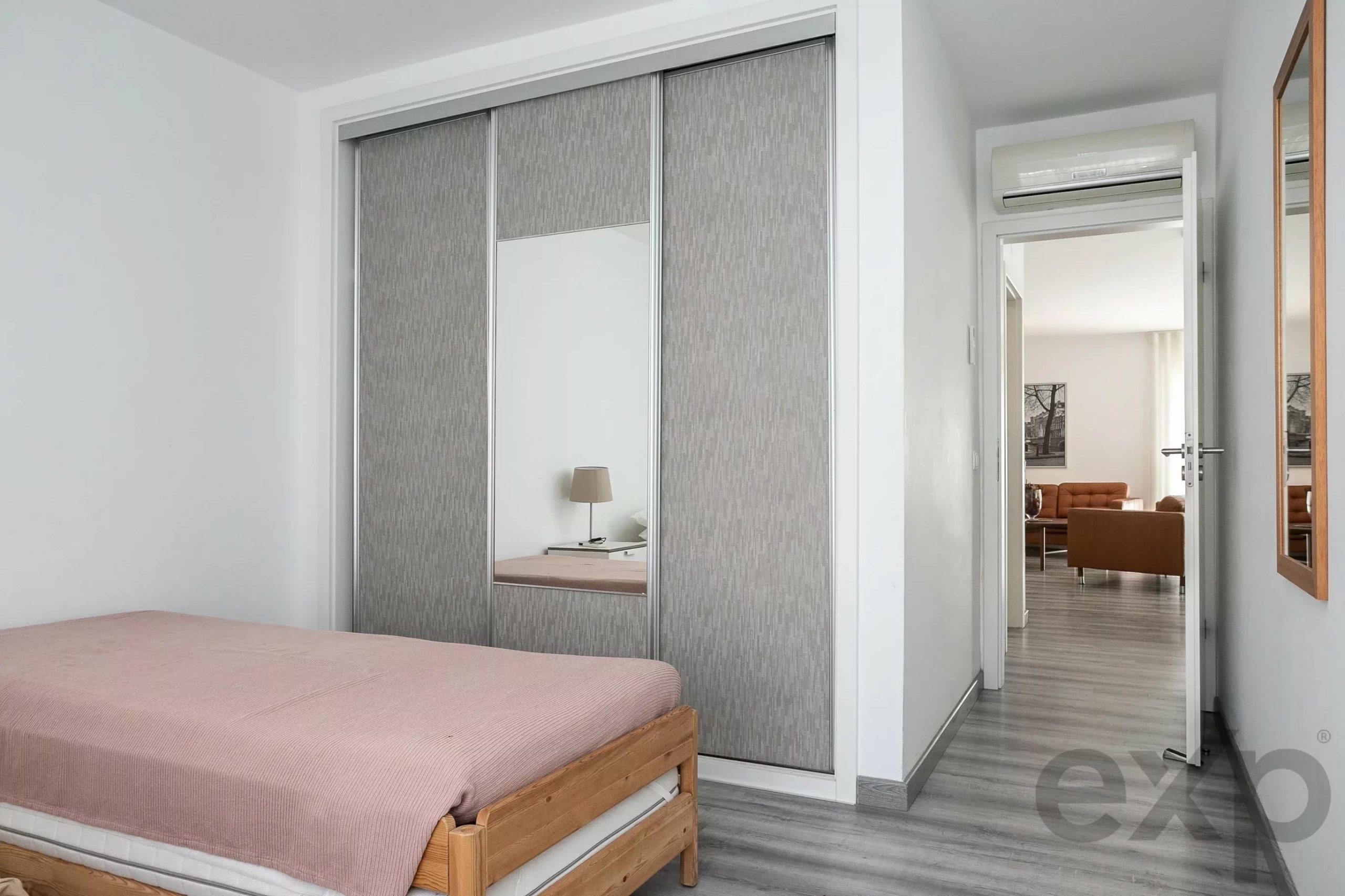 Bedroom with a wooden bed and pink bedspread, mirrored sliding closet doors, and an open door to a living room with an orange sofa.