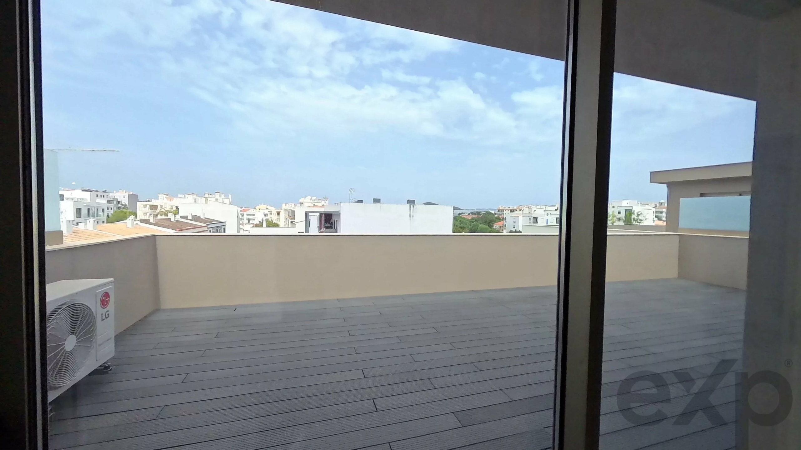 Indoor view of a glass sliding door opening to a rooftop terrace with a wooden deck and a cityscape beyond under a blue sky.