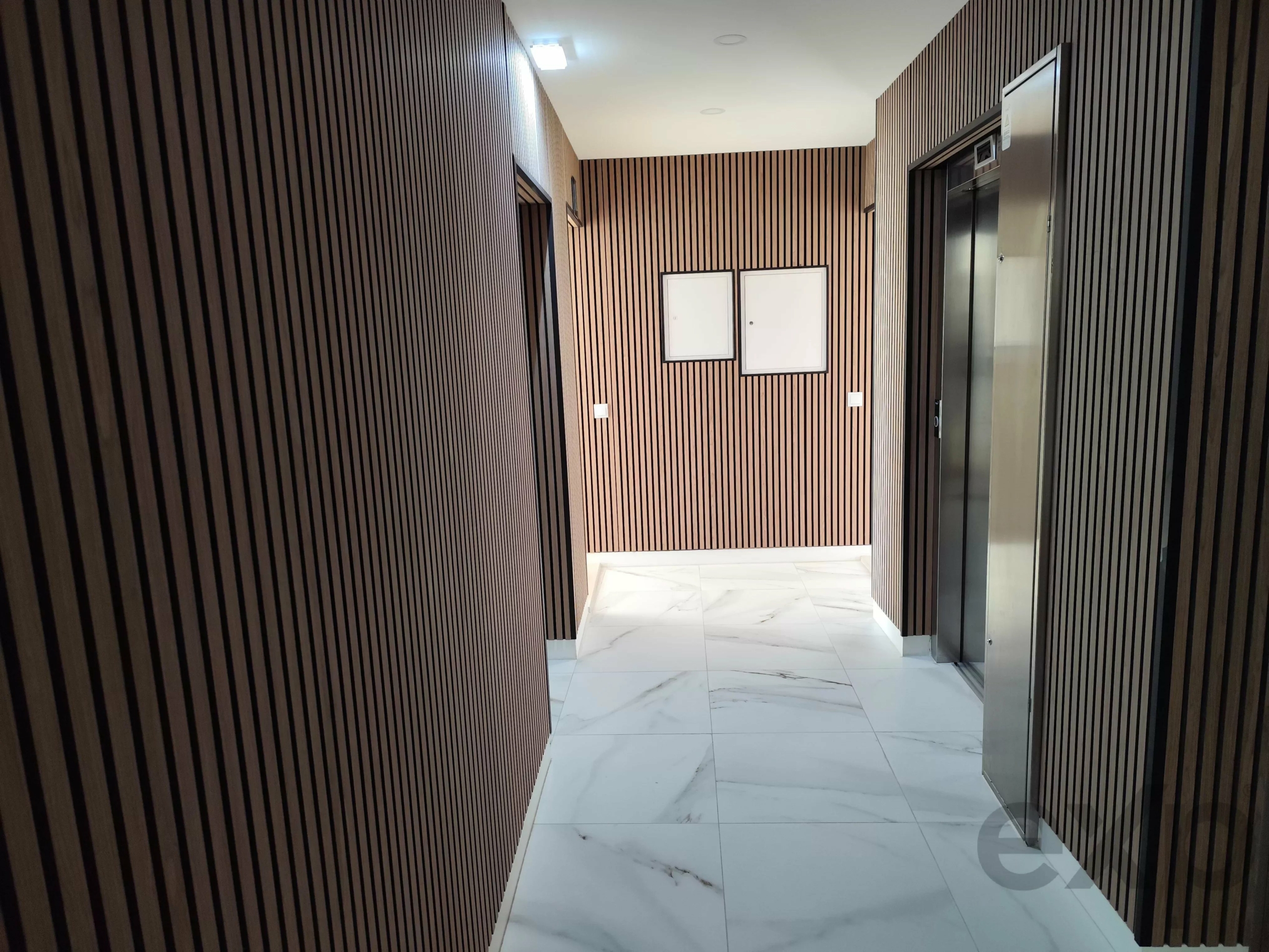 Narrow hallway with dark vertical wood slat walls, white marble floor, and an elevator on the right side.