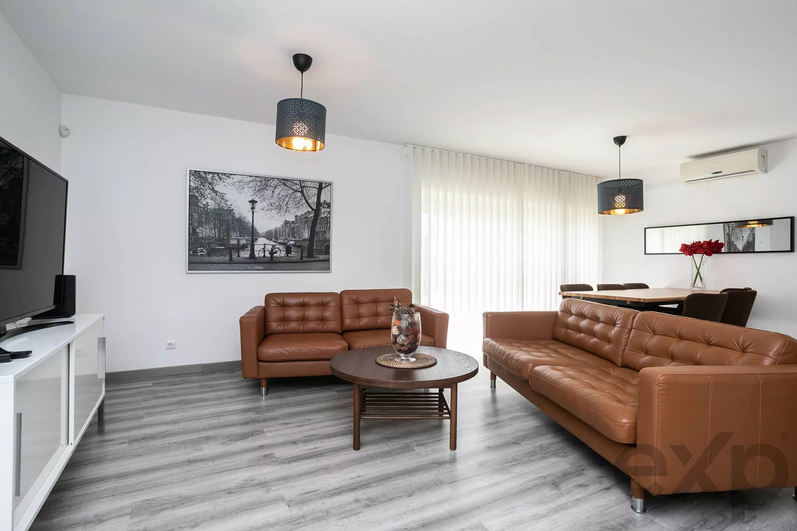 Modern living room with brown leather sofas, a round coffee table, wall art, and large windows with vertical blinds.