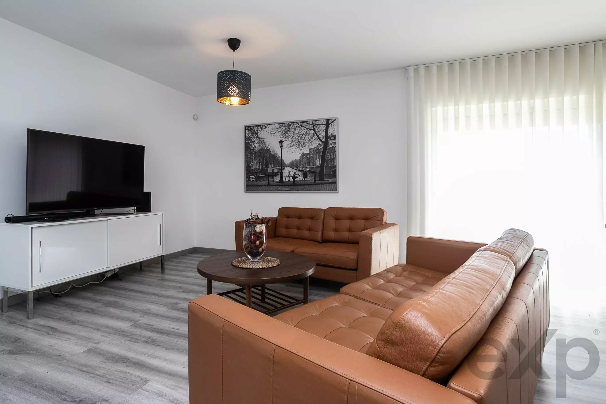 Living room with brown leather sofas, round coffee table, black-and-white cityscape wall art, and a flat-screen TV on a white cabinet.