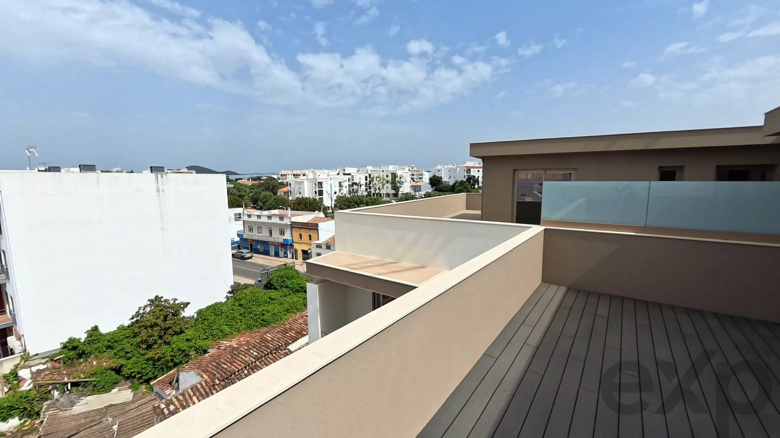 Rooftop terrace with a dark wooden deck and glass railing overlooking a white apartment neighborhood under a blue sky with clouds.