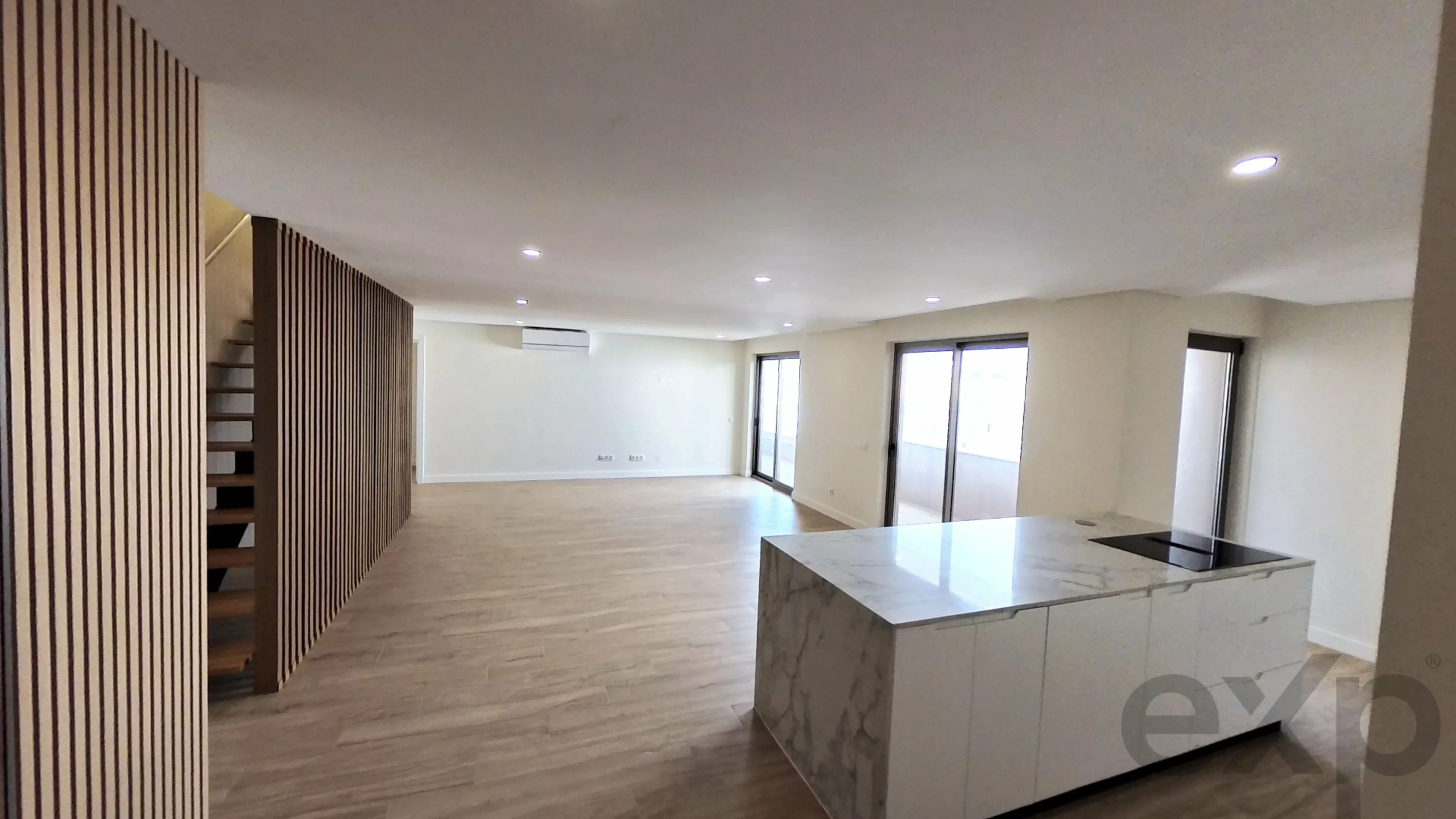 Spacious modern living area with wooden slat wall on left, staircase, large windows, and marble kitchen island with sink on the right.