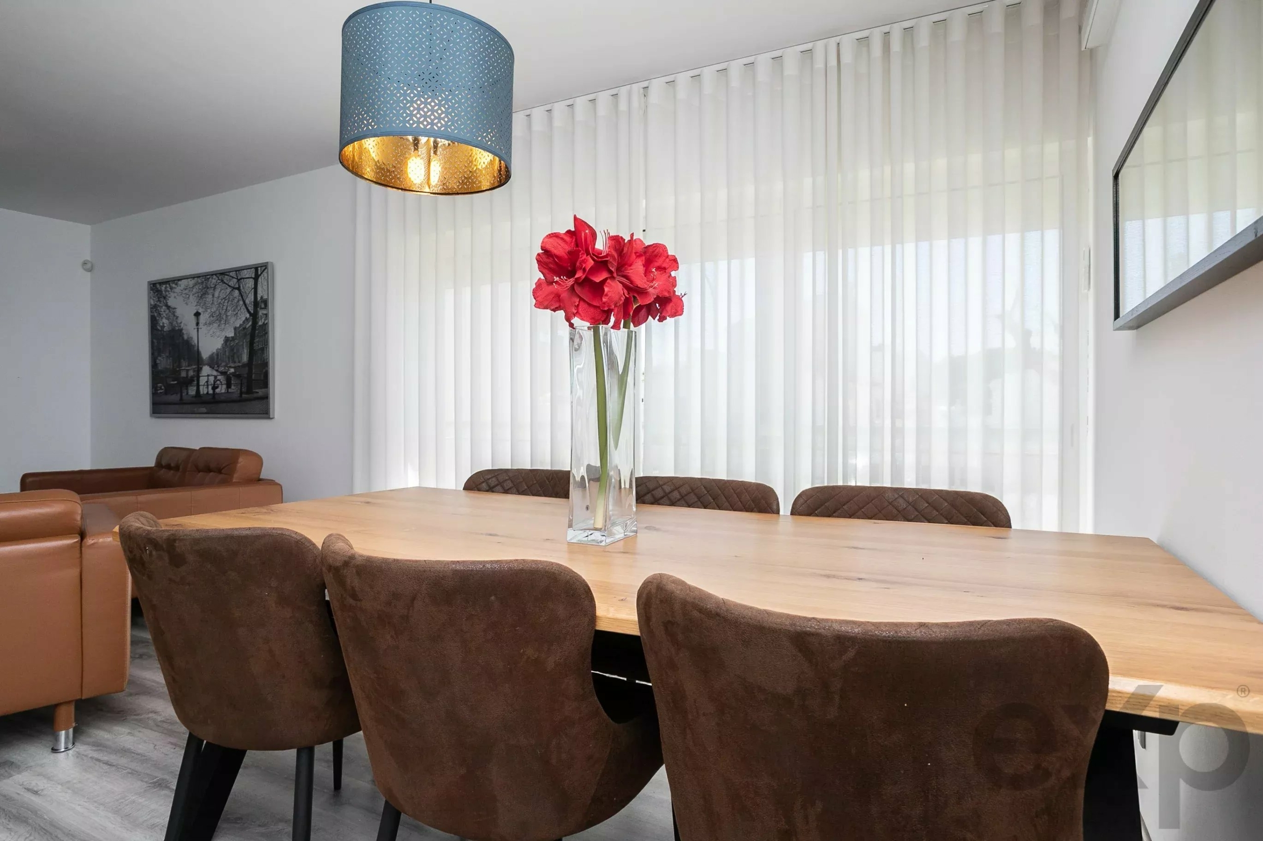 Dining room with a wooden table, brown upholstered chairs, a glass vase of red flowers, and a blue pendant light overhead.
