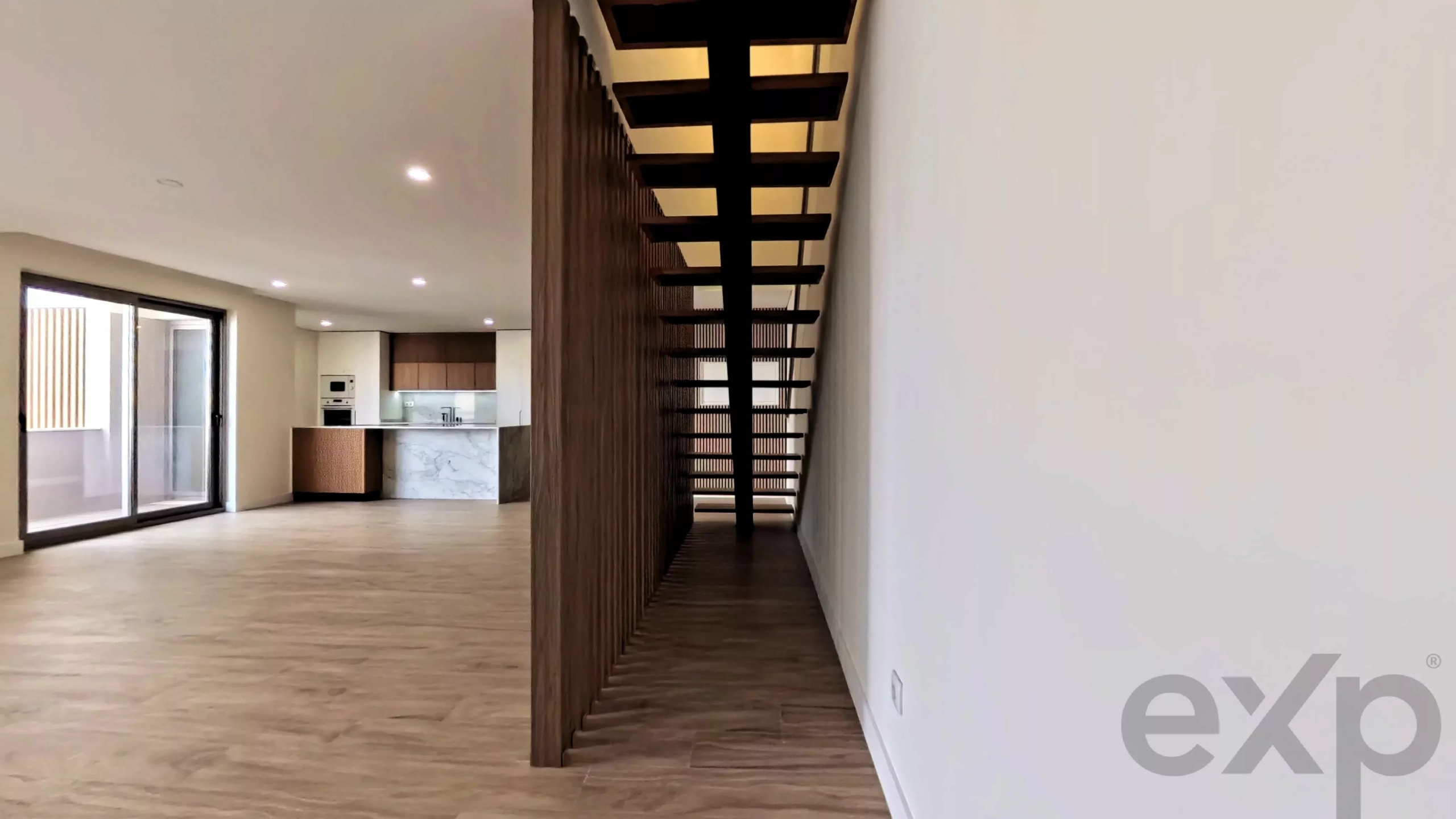 Open-concept living area with a kitchen in the back, sliding glass door on the left, and a dark open-riser staircase along a wooden divider.