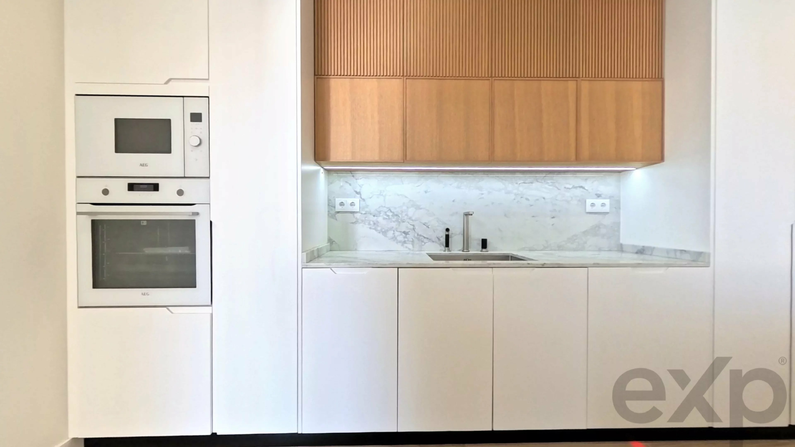Modern white kitchen with tall cabinetry, a stacked double oven on the left, and a marble backsplash with a sink and wooden upper cabinets above the counter.