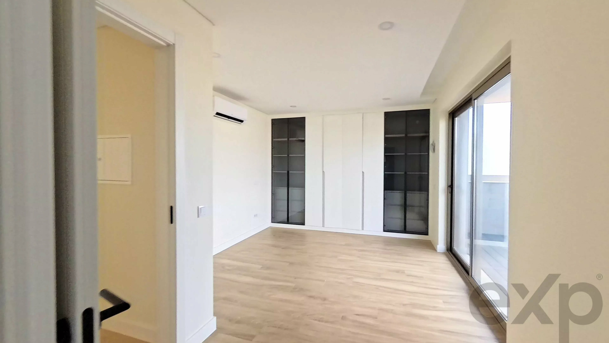 Modern empty living area with light wood flooring, white built-in cabinets with glass doors on the back wall, and sliding glass doors to a balcony on the right.