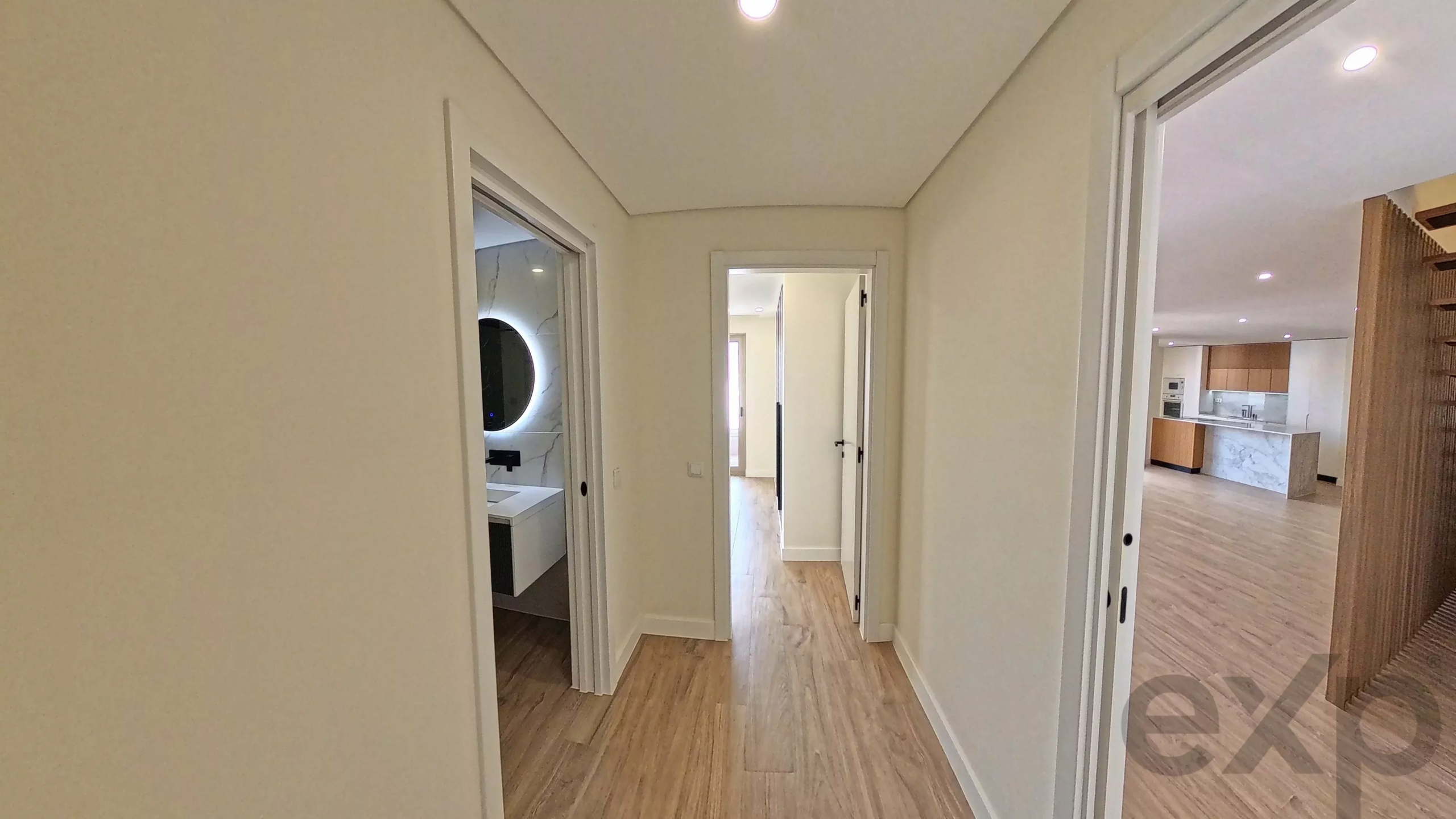 Bright hallway in a modern home with multiple doorways, leading to a bathroom with a round backlit mirror and a visible kitchen in the distance.