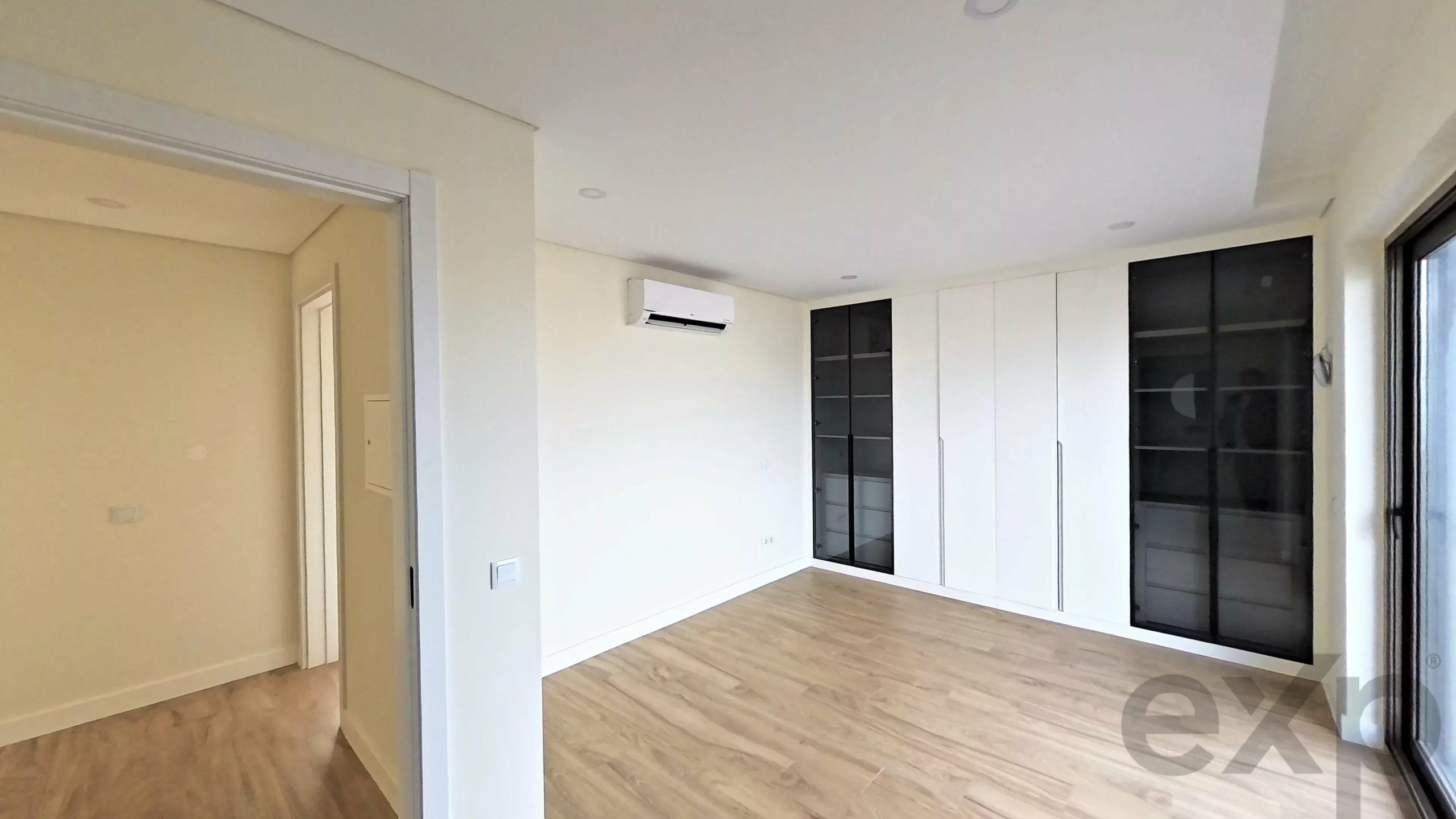 Modern empty living room with white built-in cabinets, glass-front wardrobes, wood flooring, and a wall-mounted air conditioner on the wall.