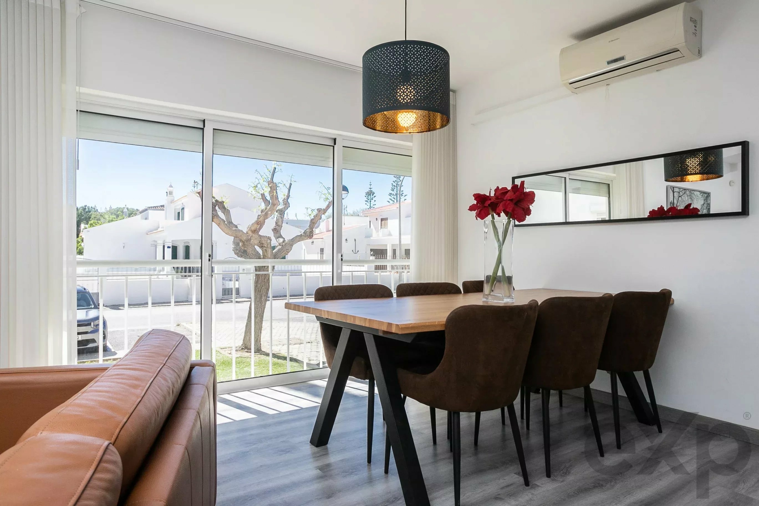 Bright dining area with a wooden table, six brown chairs, a perforated black pendant light, and a vase of red flowers by the glass doors.