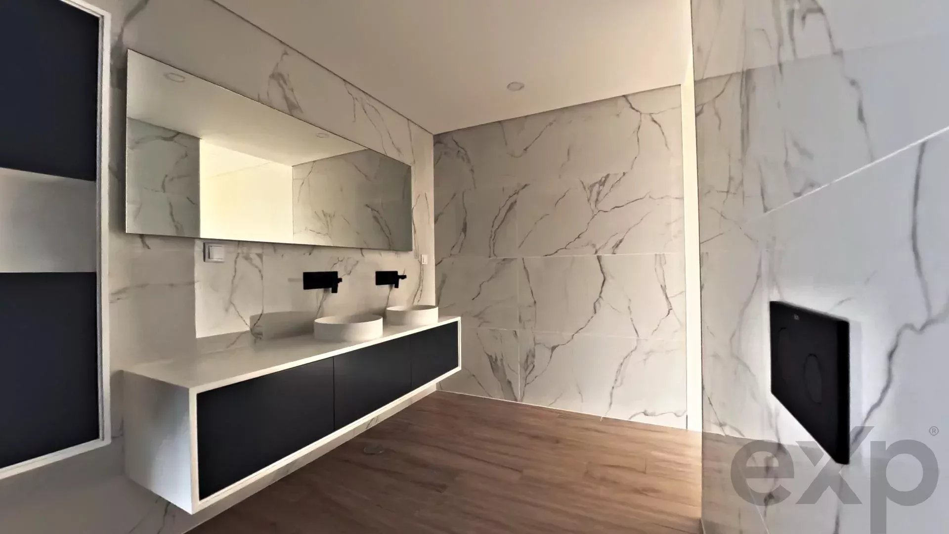 Modern bathroom with marble walls, a long white vanity with two black cabinet doors, and two white vessel sinks beneath a large mirror.