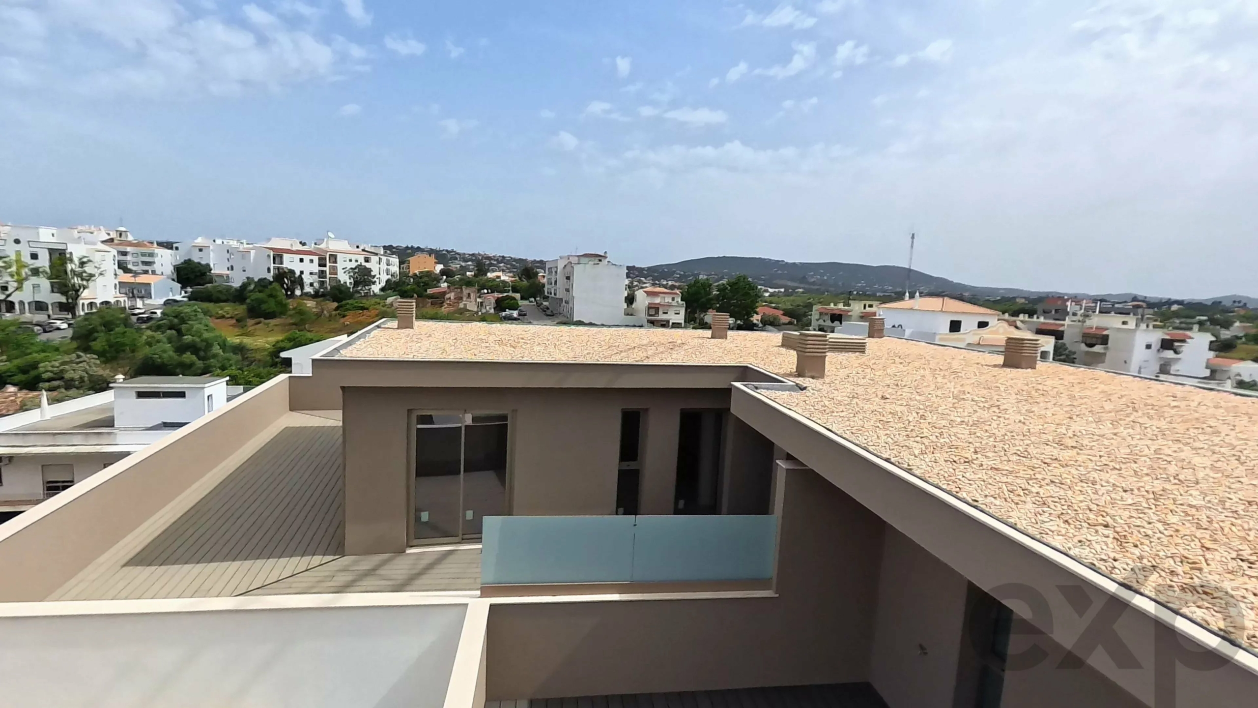 Rooftop terrace view of a residential neighborhood with white buildings and green trees under a blue sky.