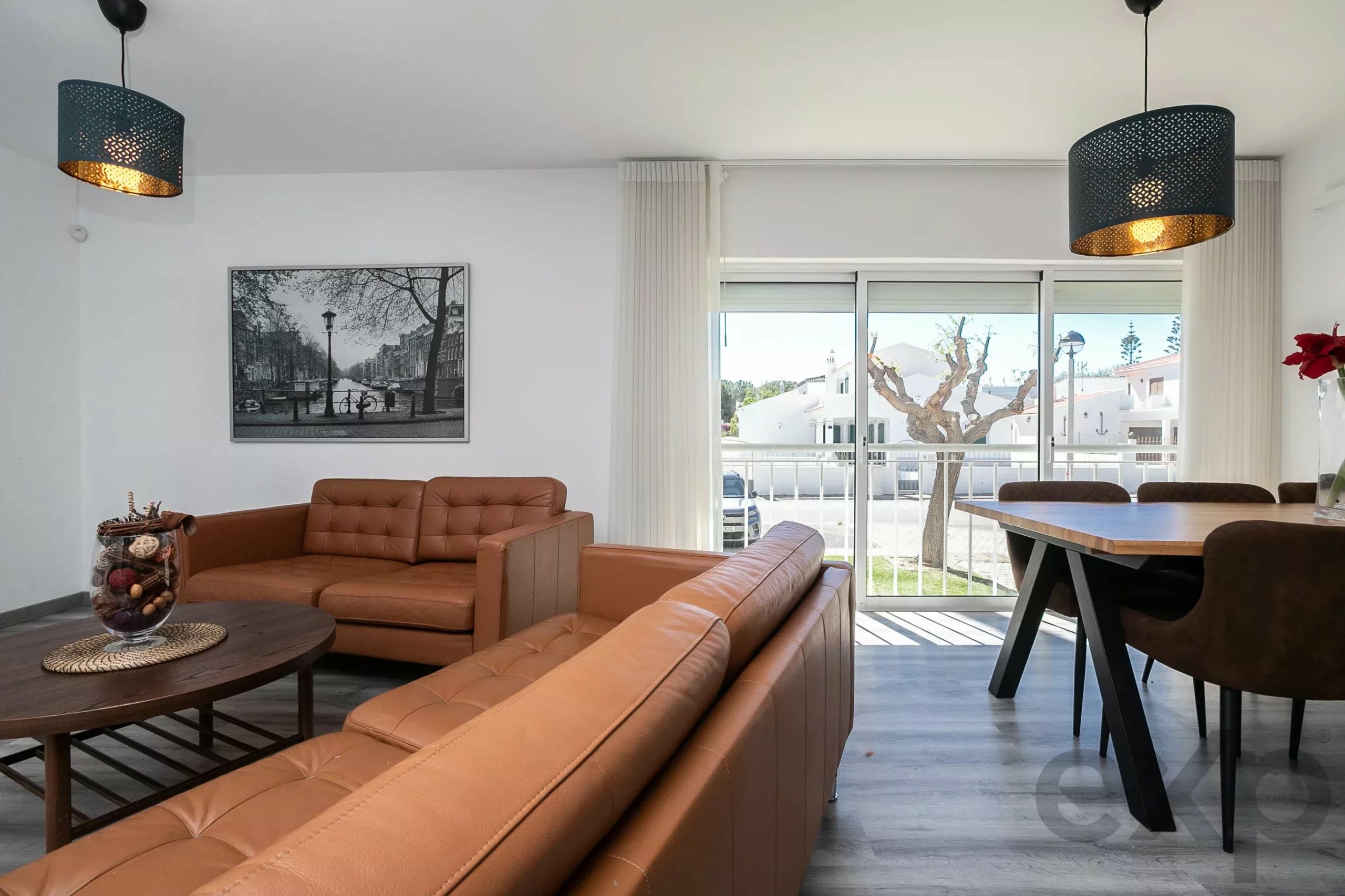 Living room with tan leather sofas, round coffee table, and a black-and-white city photo on the wall, near large glass doors with white curtains.