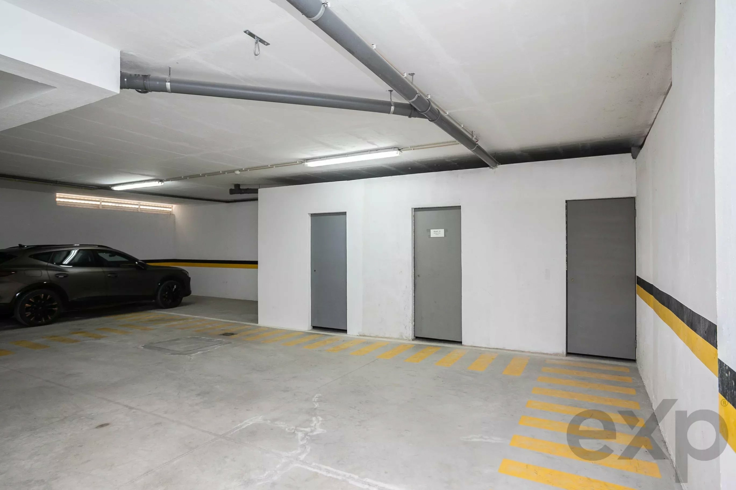 Underground parking garage with a dark gray car on the left, concrete walls, and exposed ceiling pipes with yellow striped parking lines.