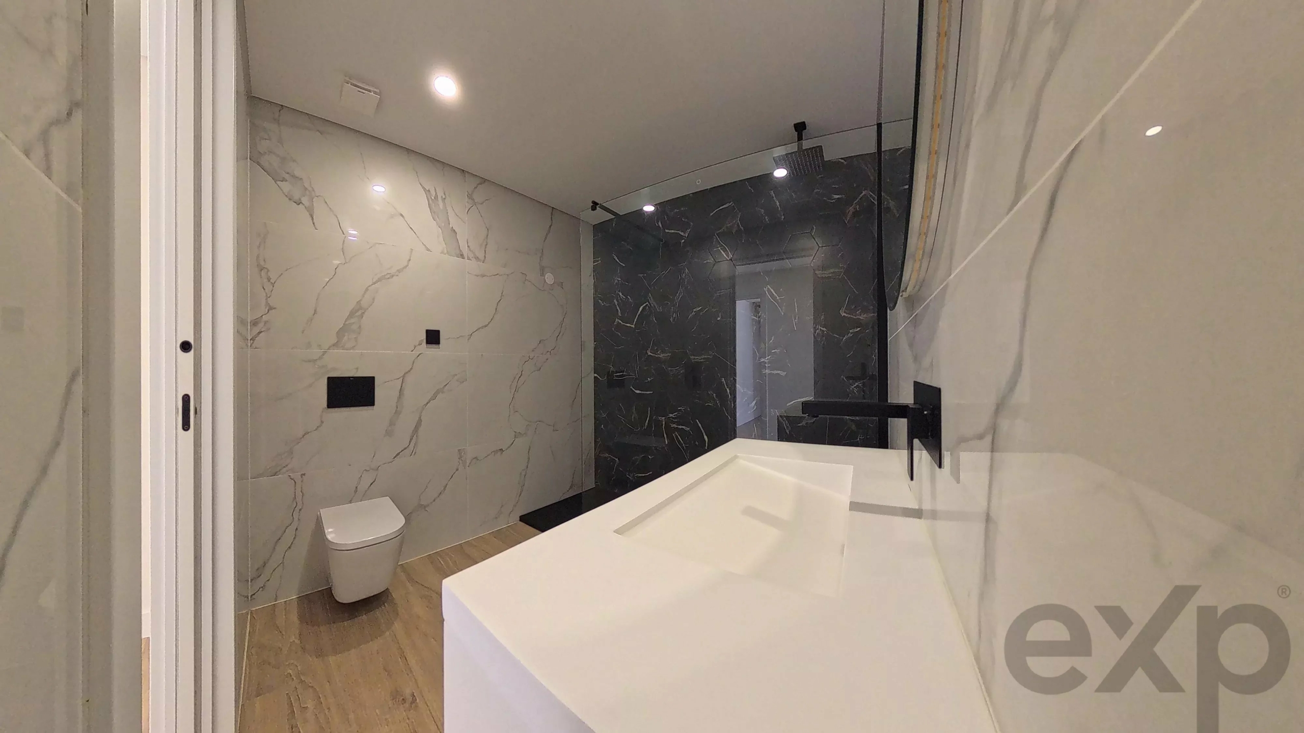 Modern bathroom with white marble walls and a dark marble accent wall, featuring a white rectangular sink and a black wall-mounted faucet. A wall-hung toilet sits nearby on wood-like flooring, with recessed ceiling lights illuminating the space.