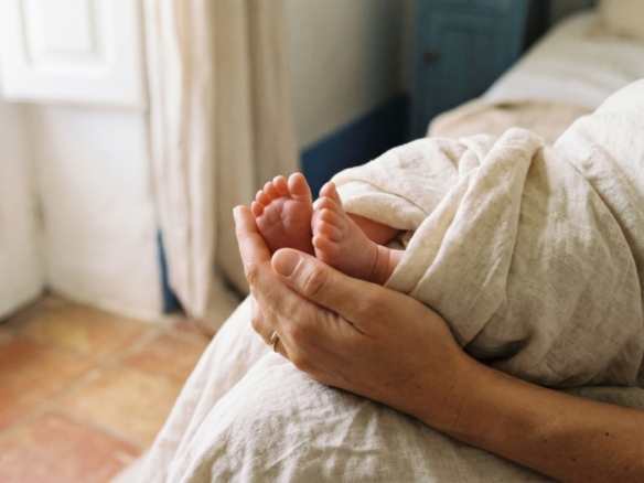 Gently cradled newborn feet held in an adult's hands, wrapped in a light blanket by a bedside scene.