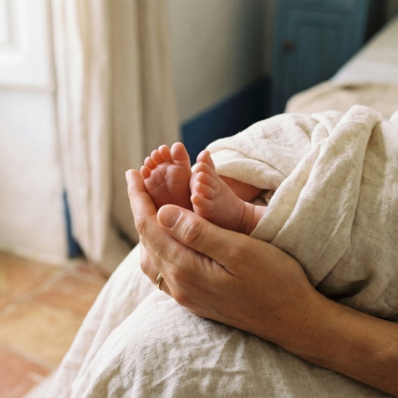 Gently cradled newborn feet held in an adult's hands, wrapped in a light blanket by a bedside scene.