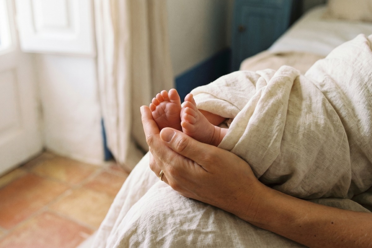 Gently cradled newborn feet held in an adult's hands, wrapped in a light blanket by a bedside scene.